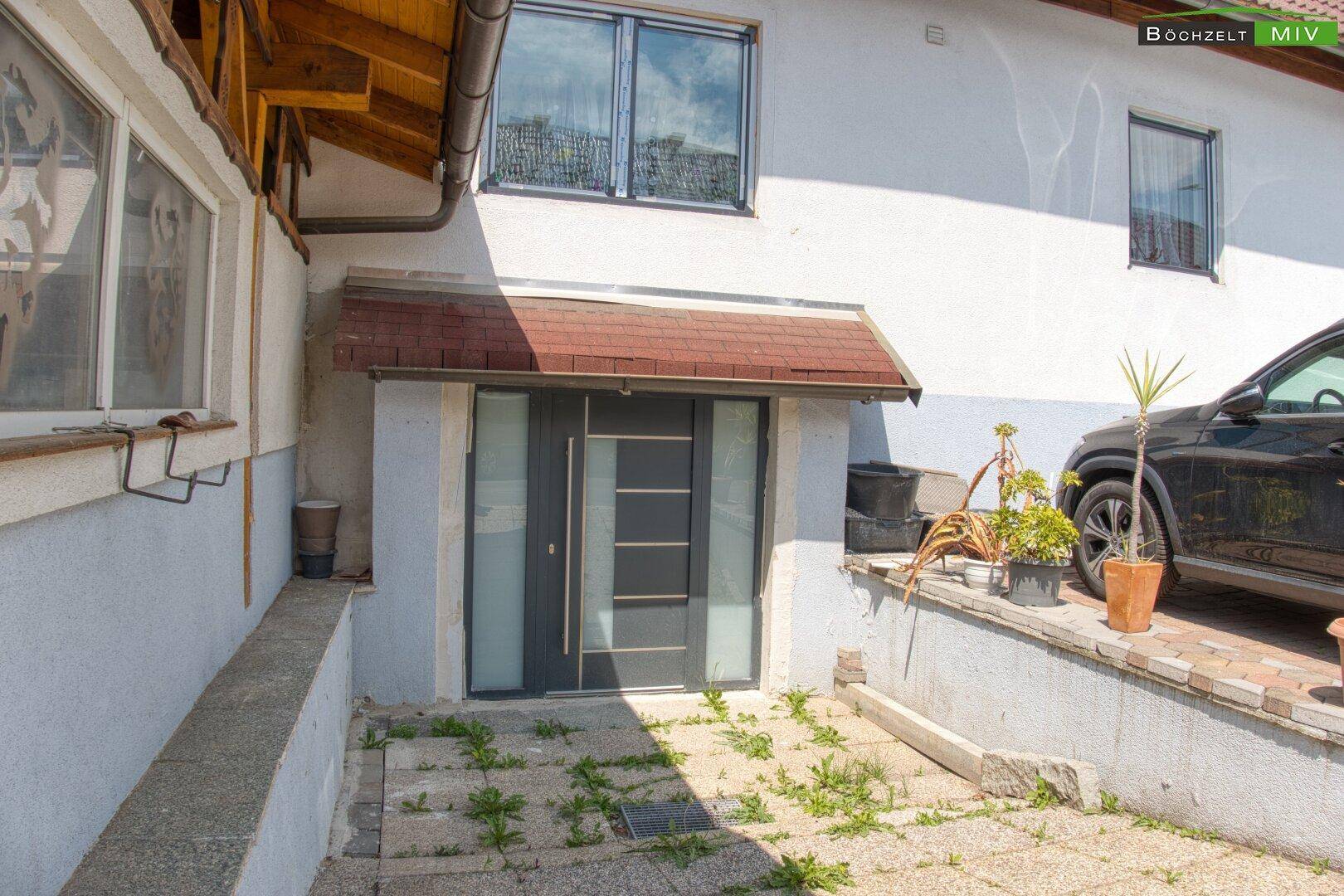 Eingangsbereich des Hauses mit moderner Haustür und einem kleinen Vordach, umgeben von Pflanzen.