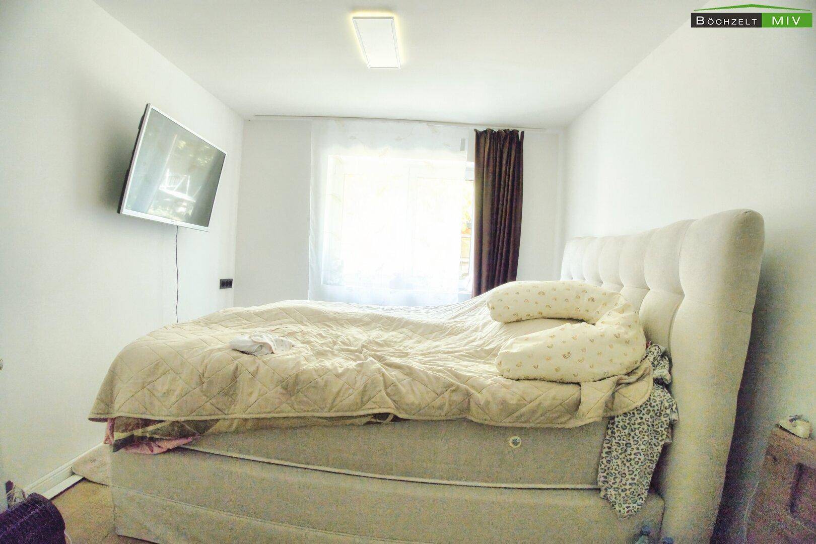 Gemütliches Schlafzimmer mit großem Bett, Fenster und an der Wand montiertem Fernseher.