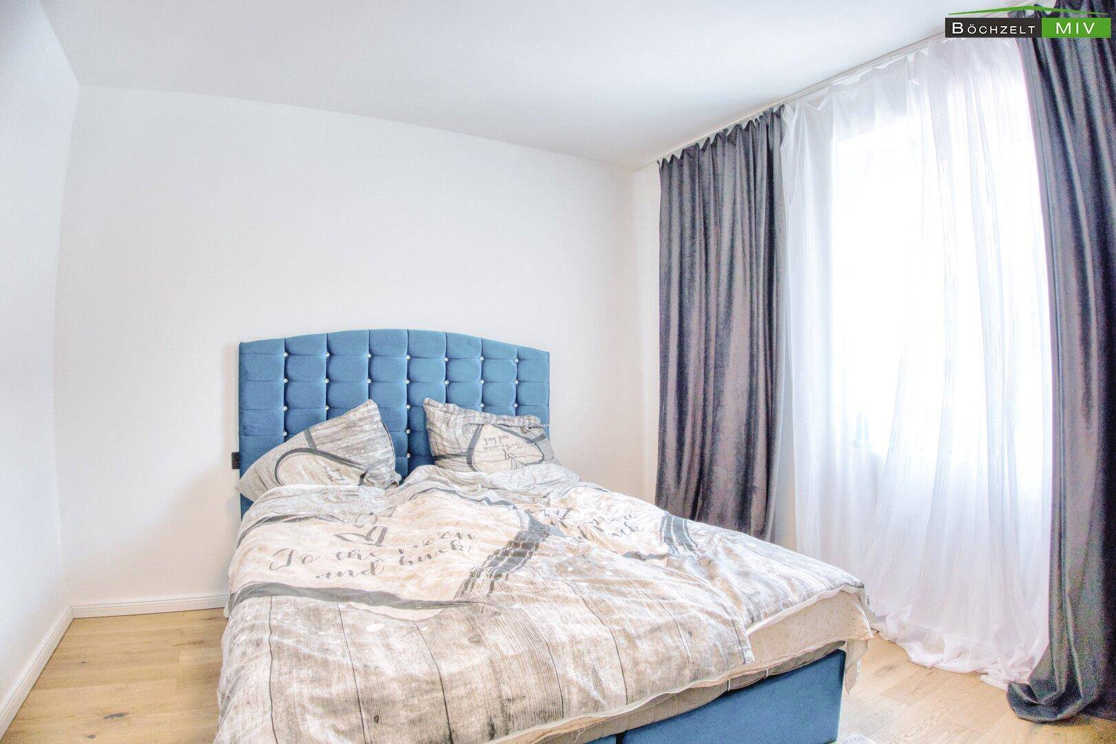 Schlafzimmer mit blauem Polsterbett, hellen Holzböden und Vorhängen.