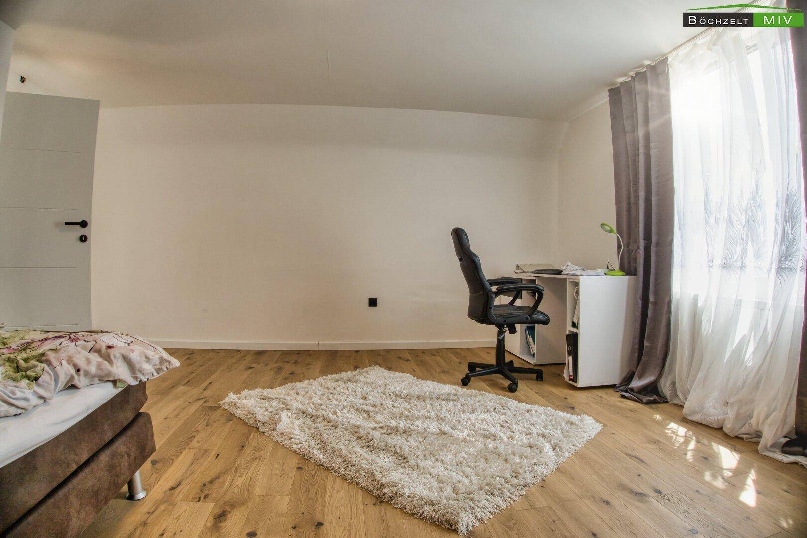 Schlafzimmer mit Holzboden, Schreibtisch und Fenster mit Vorhängen.