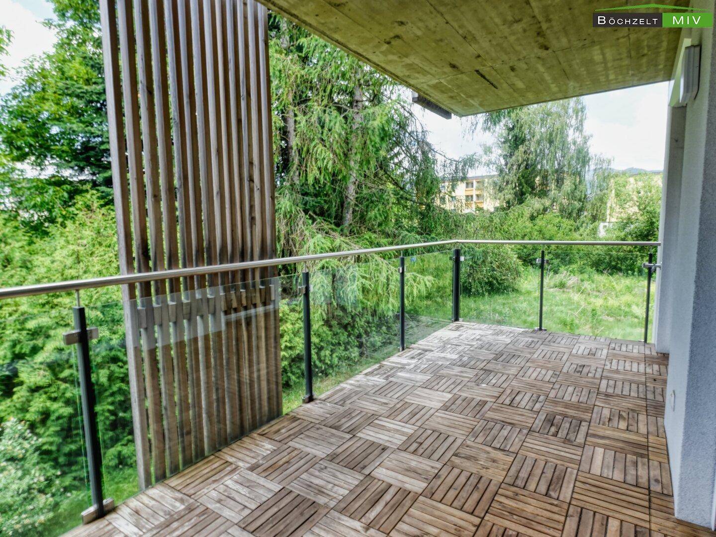 Geräumiger Balkon mit Holzbodenfliesen und Glasgeländer, umgeben von viel Grün.