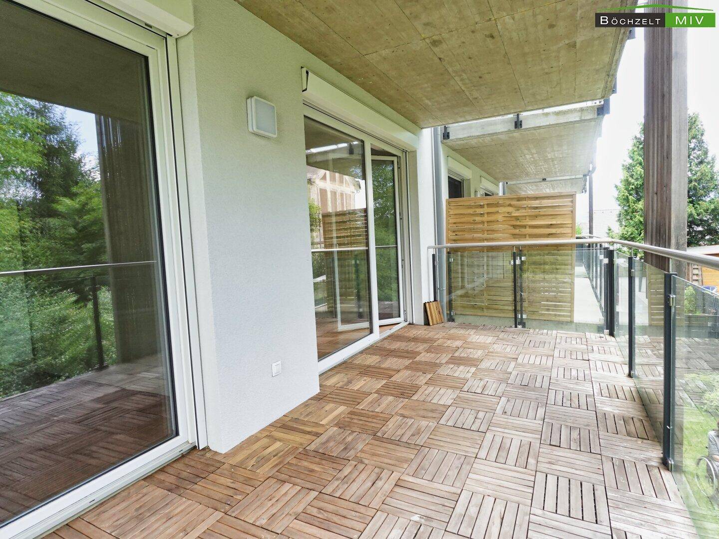 Breiter Balkon mit Holzbodenfliesen und Glasgeländer, bietet einen schönen Blick ins Grüne.