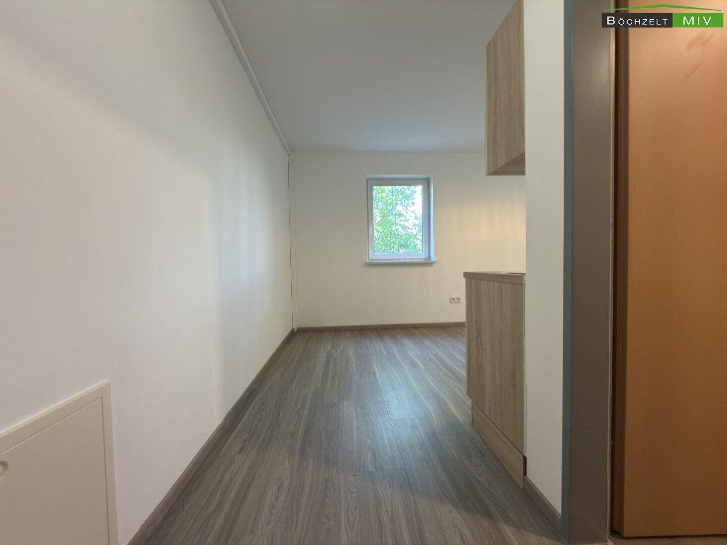 Langer, heller Raum mit Laminatboden und einem Fenster, das Tageslicht in den hinteren Bereich bringt.