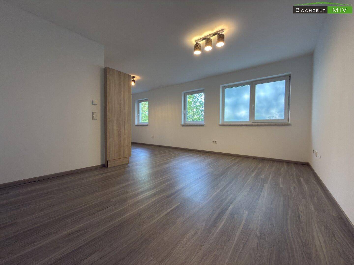 Geräumiger Wohnbereich mit hellem Laminatboden, weißen Wänden und mehreren Fenstern für viel Tageslicht.