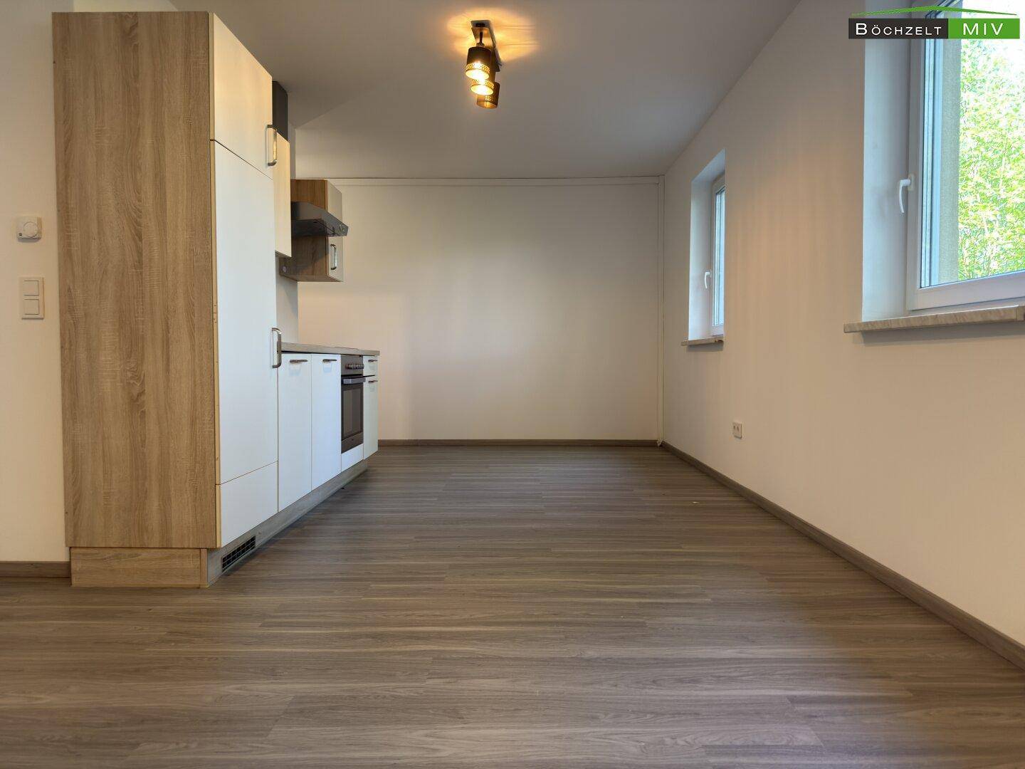 Geräumiger Wohnbereich mit integrierter Küchenzeile, hellem Laminatboden und großen Fenstern mit Blick ins Grüne.