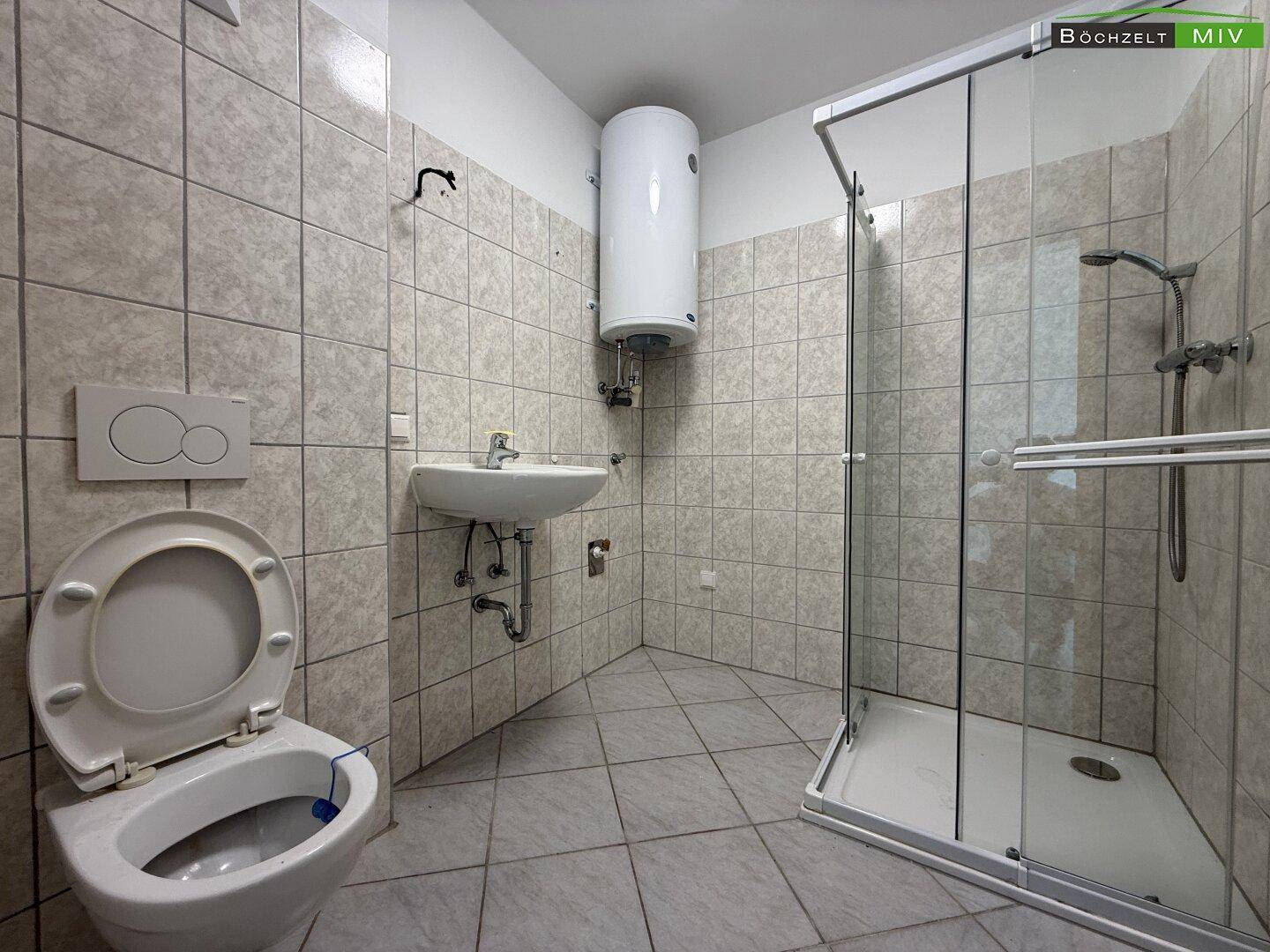 Gefliestes Badezimmer mit Toilette, Waschbecken, Duschkabine und Warmwasserboiler an der Wand.