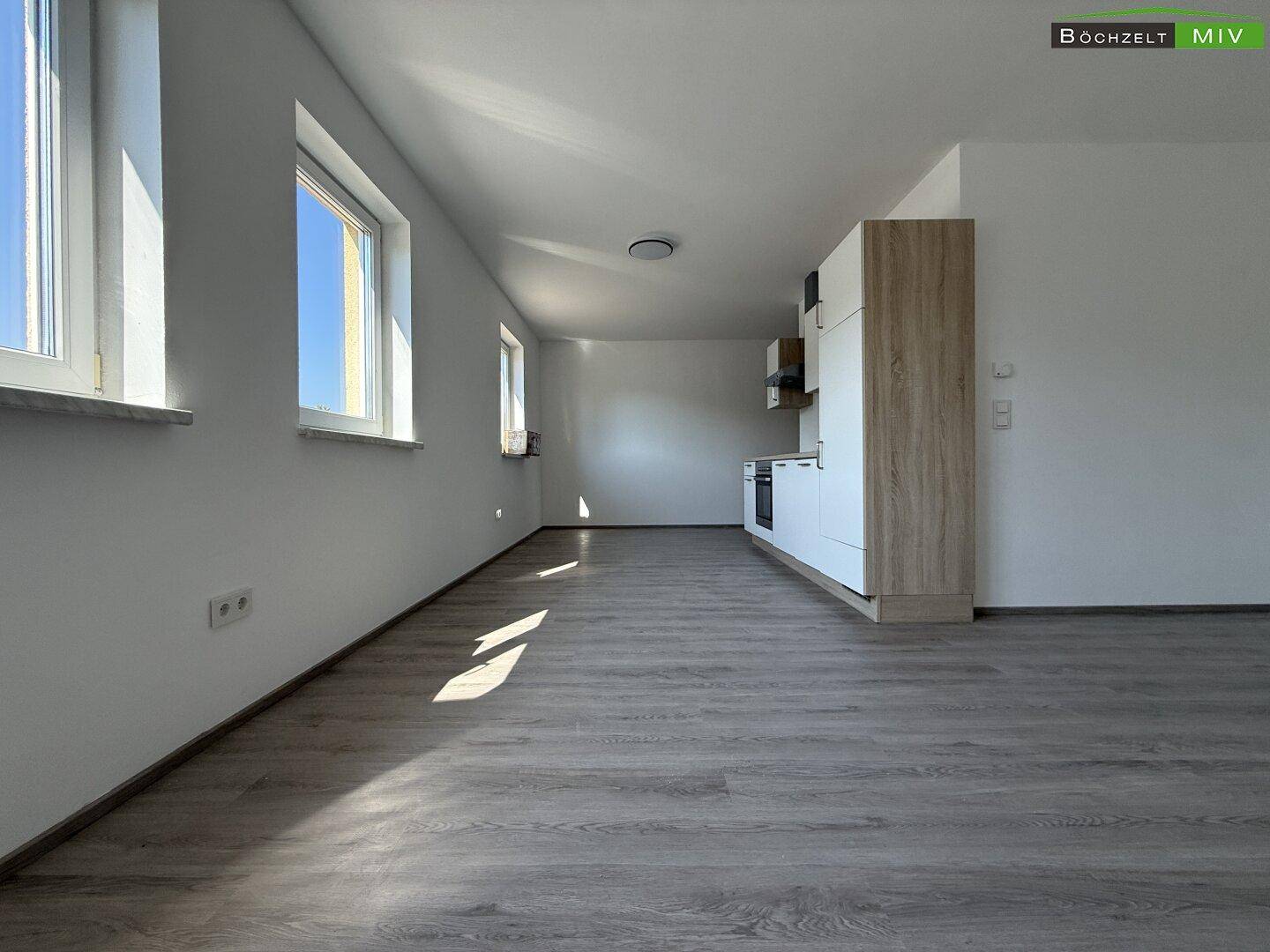 Heller, offener Raum mit Küchenzeile, Laminatboden und mehreren Fenstern, die viel Sonnenlicht hereinlassen.