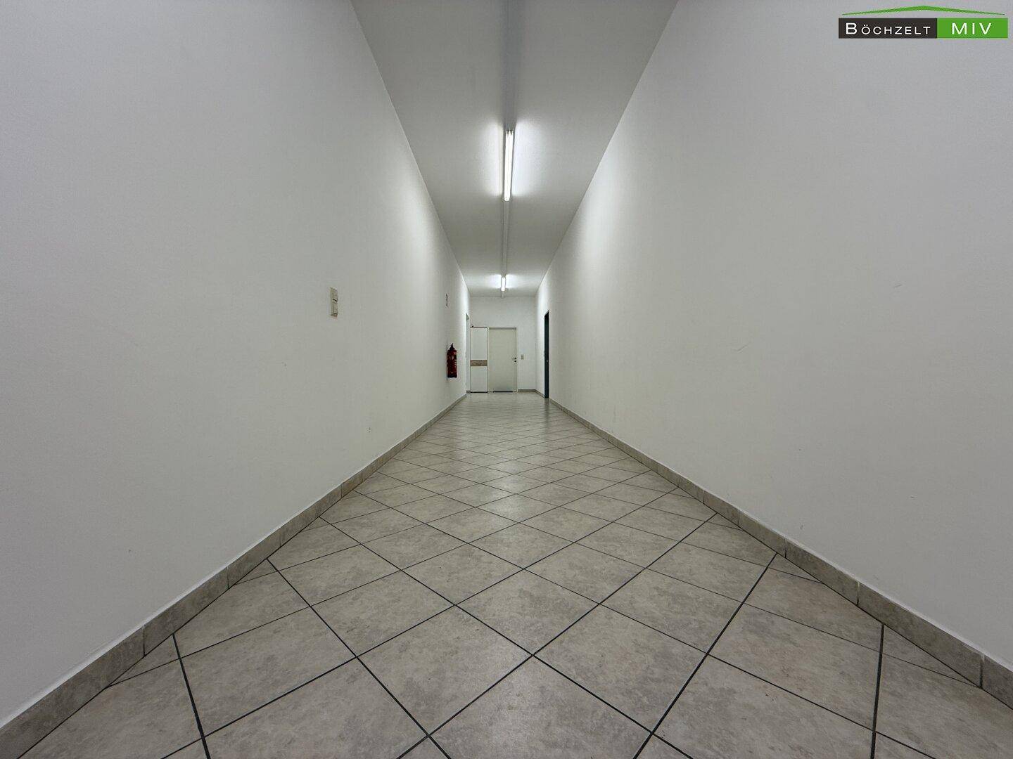 Langer, gefliester Flur mit weißen Wänden und Deckenbeleuchtung, der zu weiteren Räumen führt.