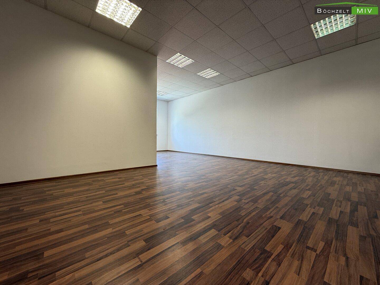 Geräumiger, leerer Raum mit dunklem Laminatboden und Rasterdeckenplatten, ideal für Büronutzung.