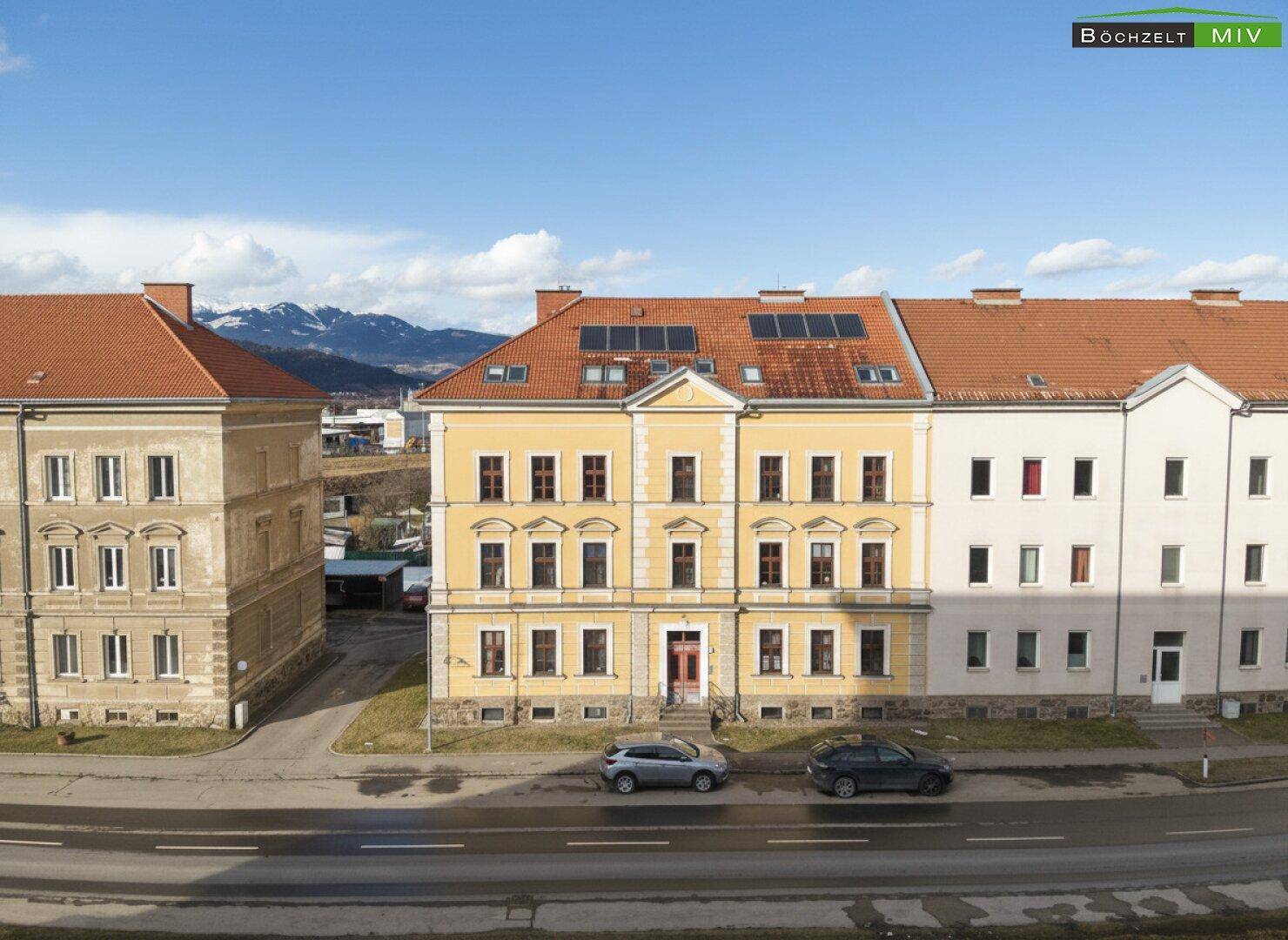 Ansicht eines historischen Gebäudes mit gelber Fassade und roten Dächern vor einer Bergkulisse.