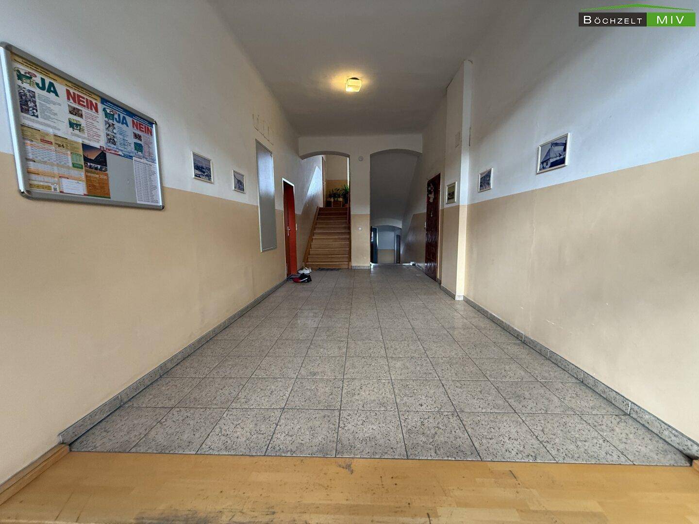Langer Flur im Eingangsbereich eines Gebäudes mit Fliesenboden und einer Treppe im Hintergrund.