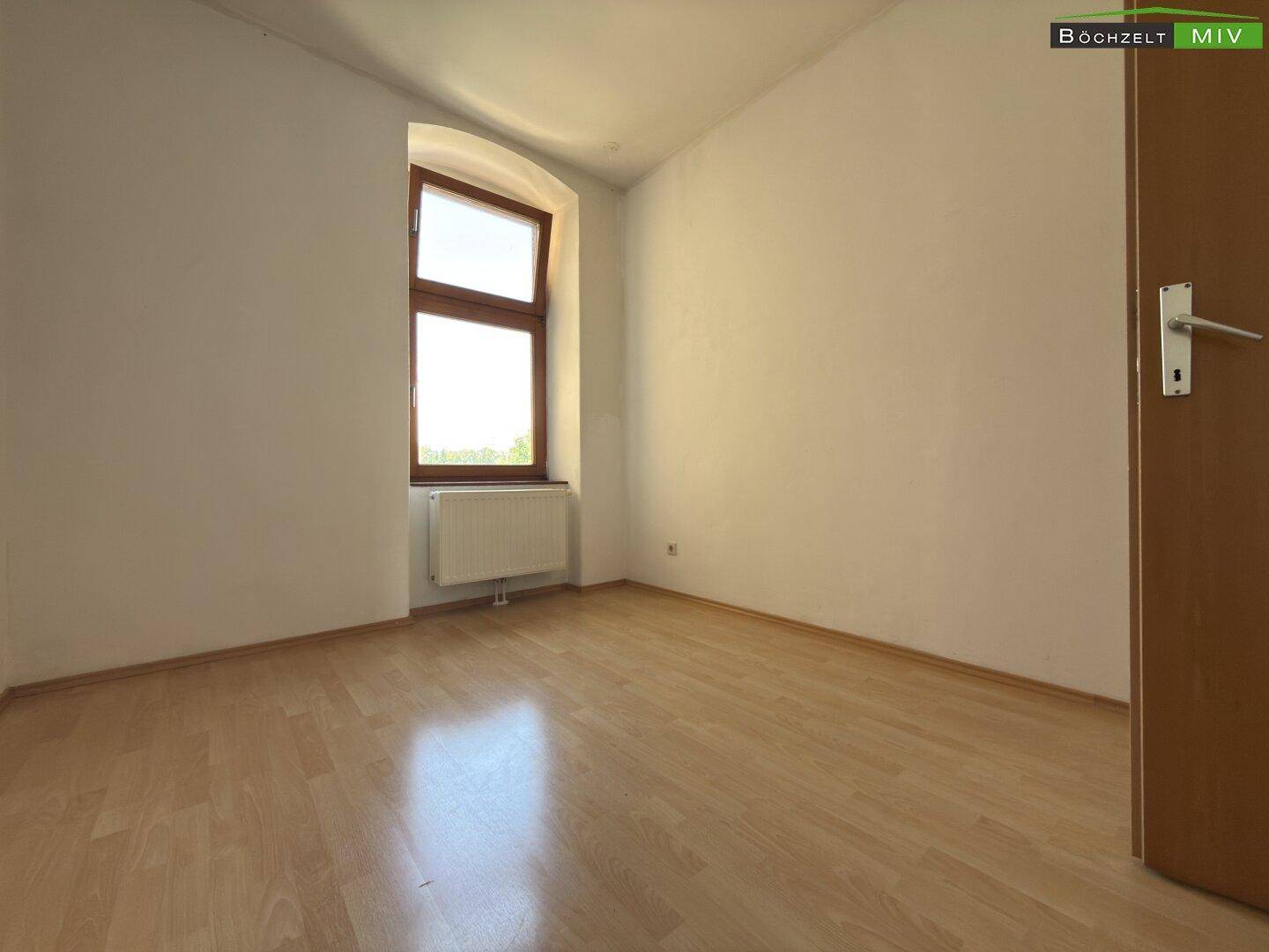 Helles Zimmer mit Laminatboden, weißen Wänden und einem Fenster mit Blick ins Grüne.