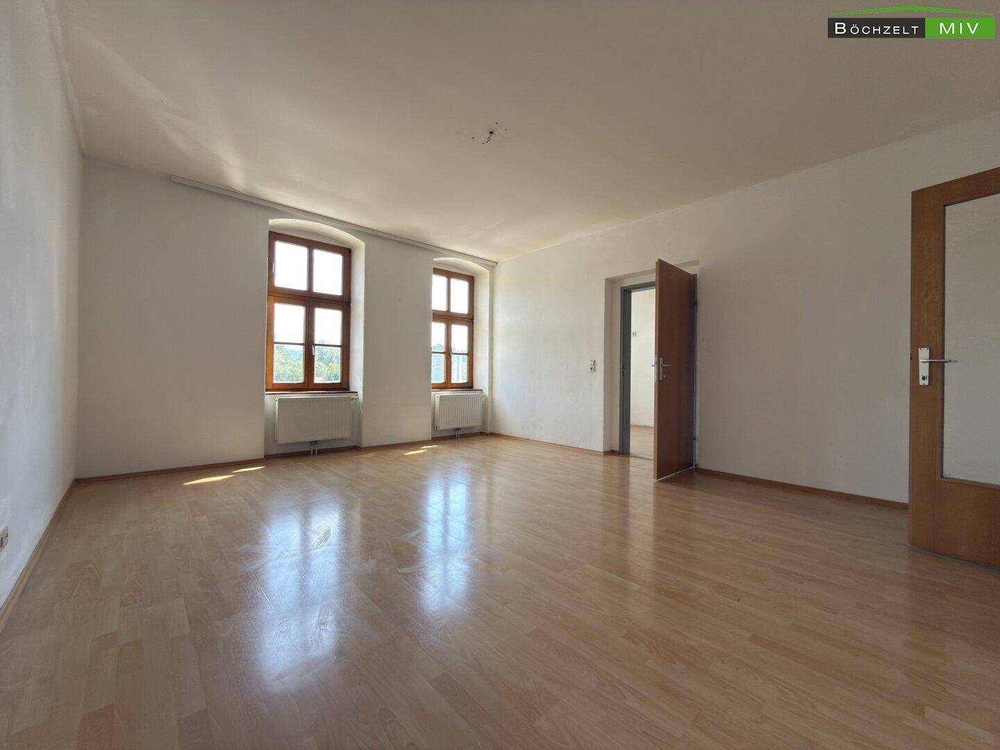 Großes, helles Zimmer mit Laminatboden und zwei Fenstern, ideal für einen Wohnbereich.