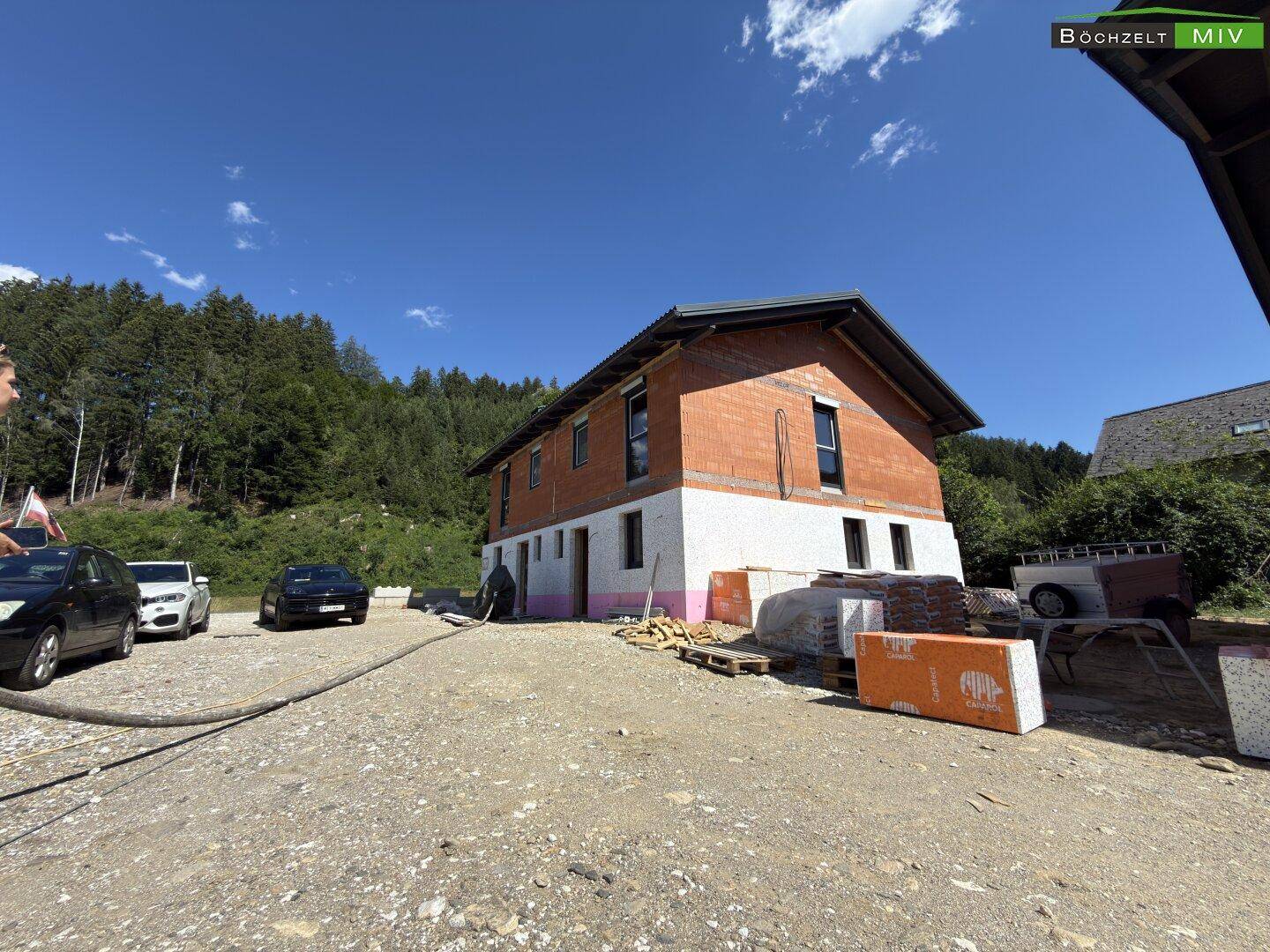 Weitläufige Ansicht des im Bau befindlichen Hauses mit Ziegelmauerwerk und Dämmung vor bewaldeter Hügellandschaft.