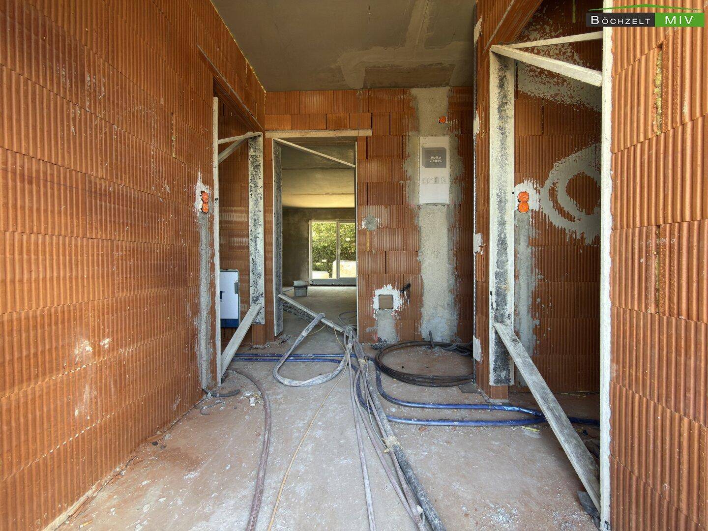 Flur im Rohbau mit unverputzten Ziegelwänden und sichtbaren Elektro- und Sanitärinstallationen auf dem Boden.