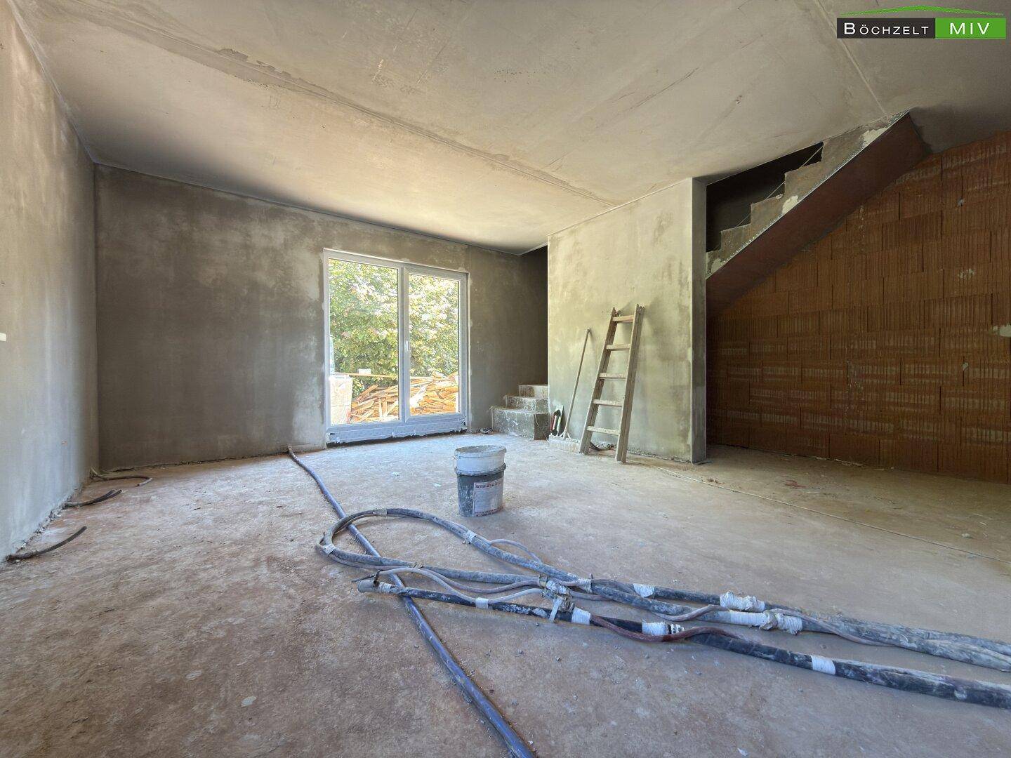 Geräumiger Wohnbereich im Rohbau mit großen Fenstern, die viel Tageslicht hereinlassen, und Treppenansatz.