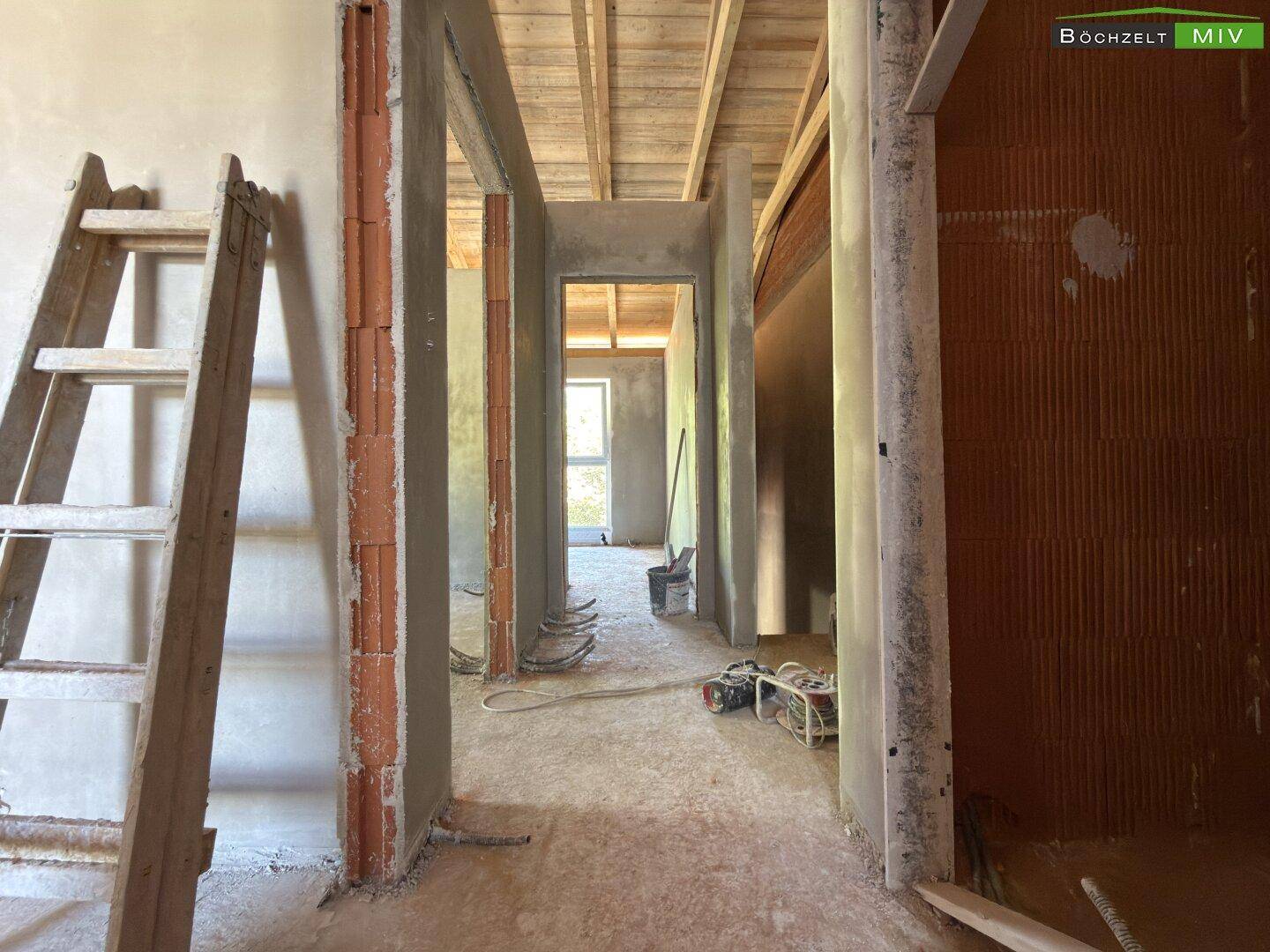 Flur im Obergeschoss des Rohbaus mit sichtbarer Holzkonstruktion des Daches und verputzten Wänden.