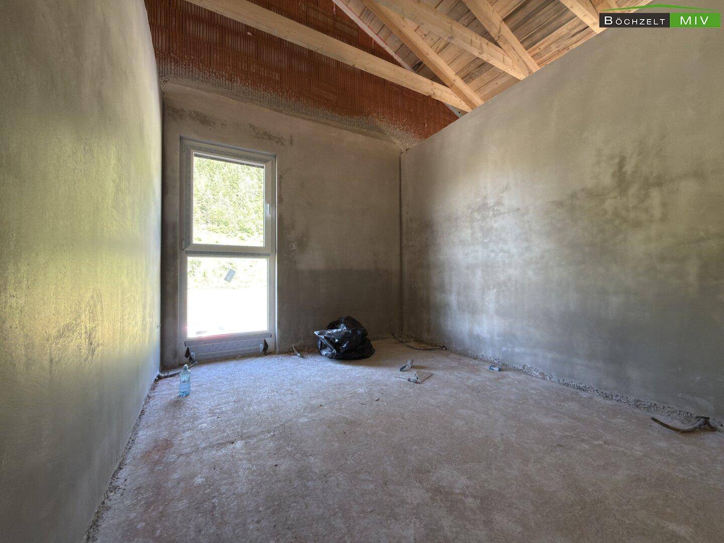 Zimmer im Dachgeschoss mit schrägen Wänden, sichtbarer Holzdecke und einem Fenster für natürliches Licht.