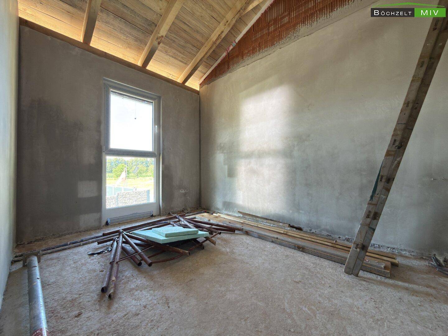Dachgeschosszimmer im Rohbau mit schrägen Wänden, sichtbarer Holzkonstruktion und Baumaterialien auf dem Boden.