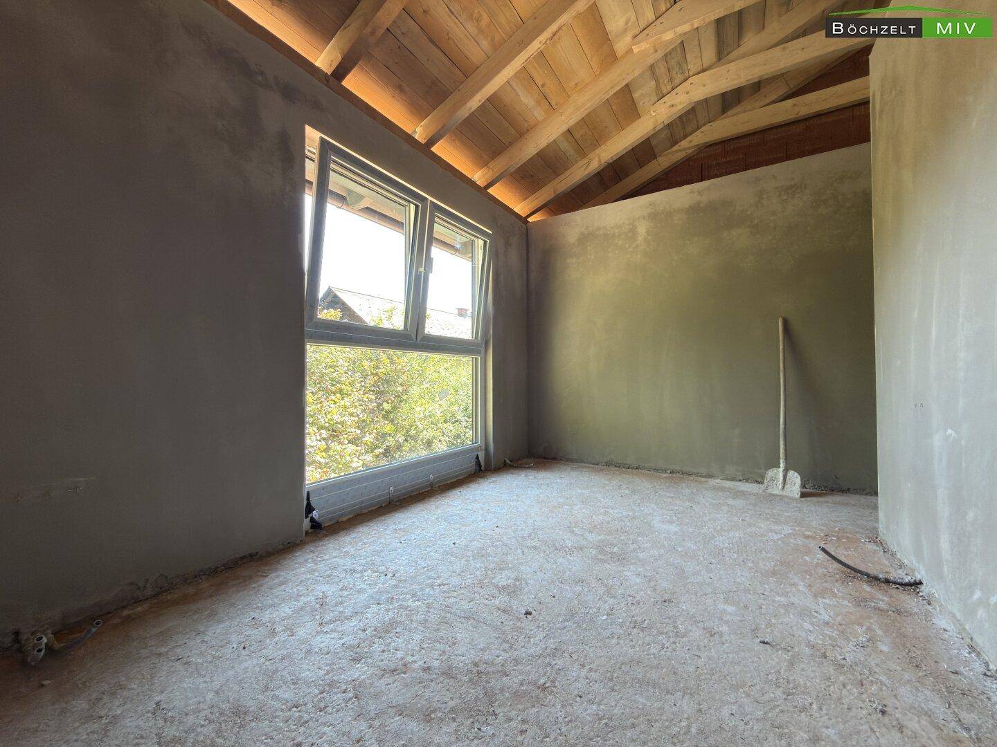 Helles Dachgeschosszimmer im Rohbau mit großem Fenster, schrägen Wänden und sichtbarer Holzdecke.