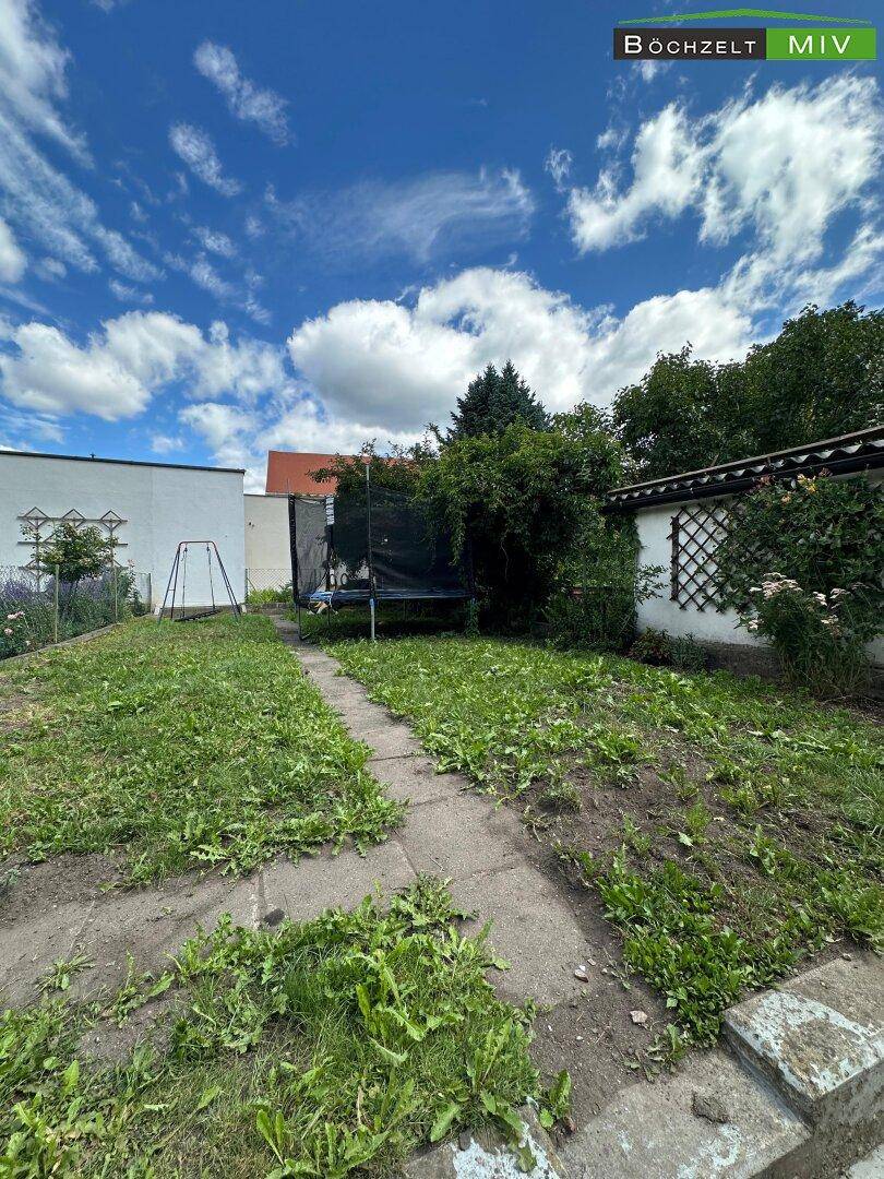 Gartenbereich mit Rasen, Trampolin und Bäumen im Hintergrund unter blauem Himmel.