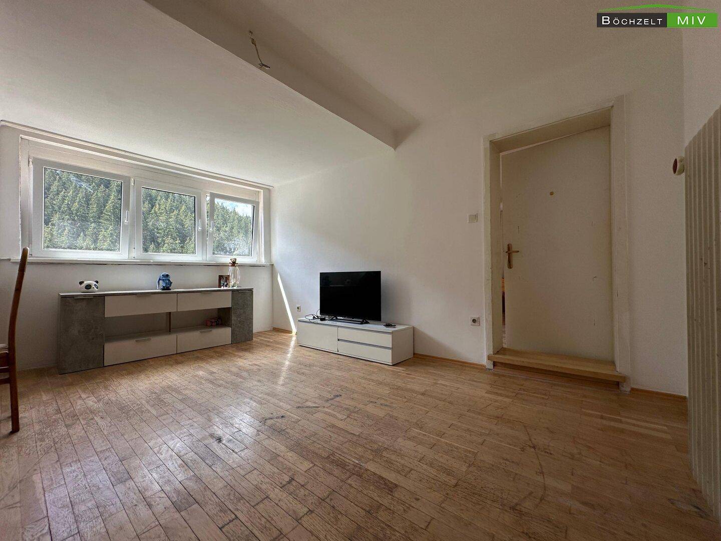 Heller Wohnraum mit Holzboden, Fernseher und einem großen Fensterbereich.