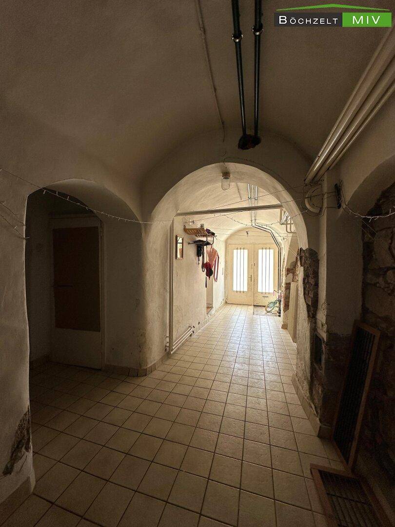 Gewölbter Kellerflur mit gefliestem Boden und Blick auf eine Tür am Ende des Ganges.