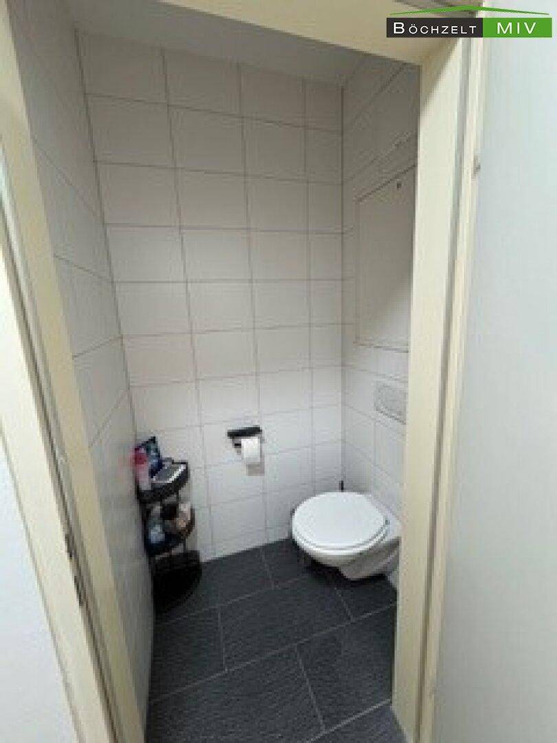 Separates WC mit wandhängender Toilette und dunklen Bodenfliesen.