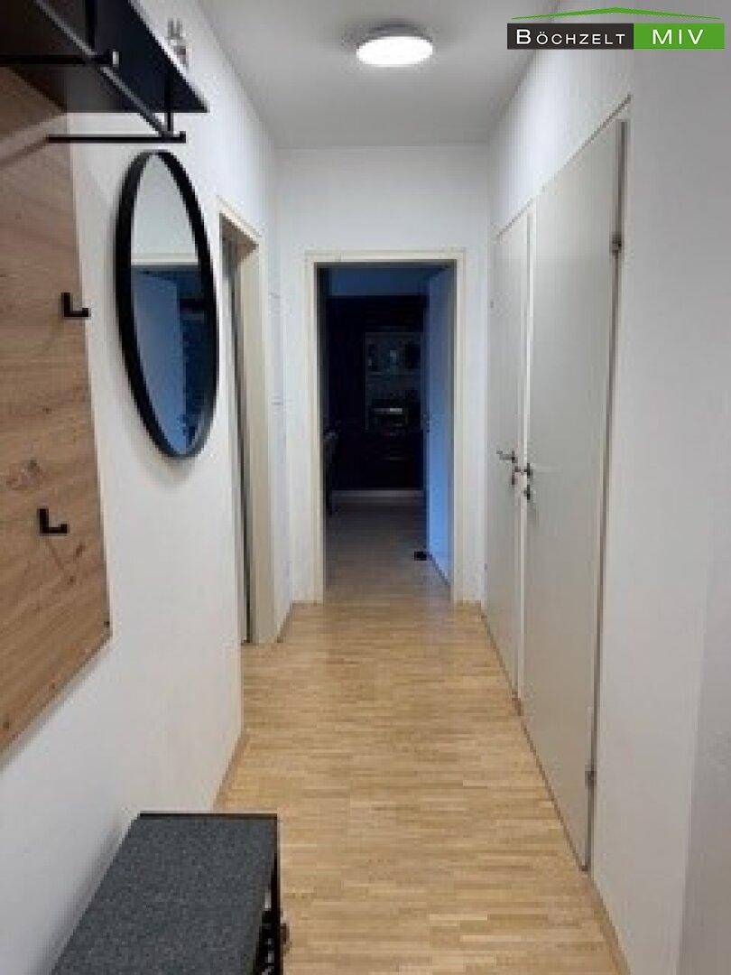 Heller Flur mit Holzboden, ovalem Spiegel und Garderobe, der zu weiteren Räumen führt.