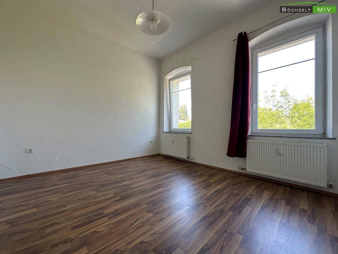 Helles Schlafzimmer mit Laminatboden, weißen Wänden und zwei Fenstern mit Blick ins Grüne.