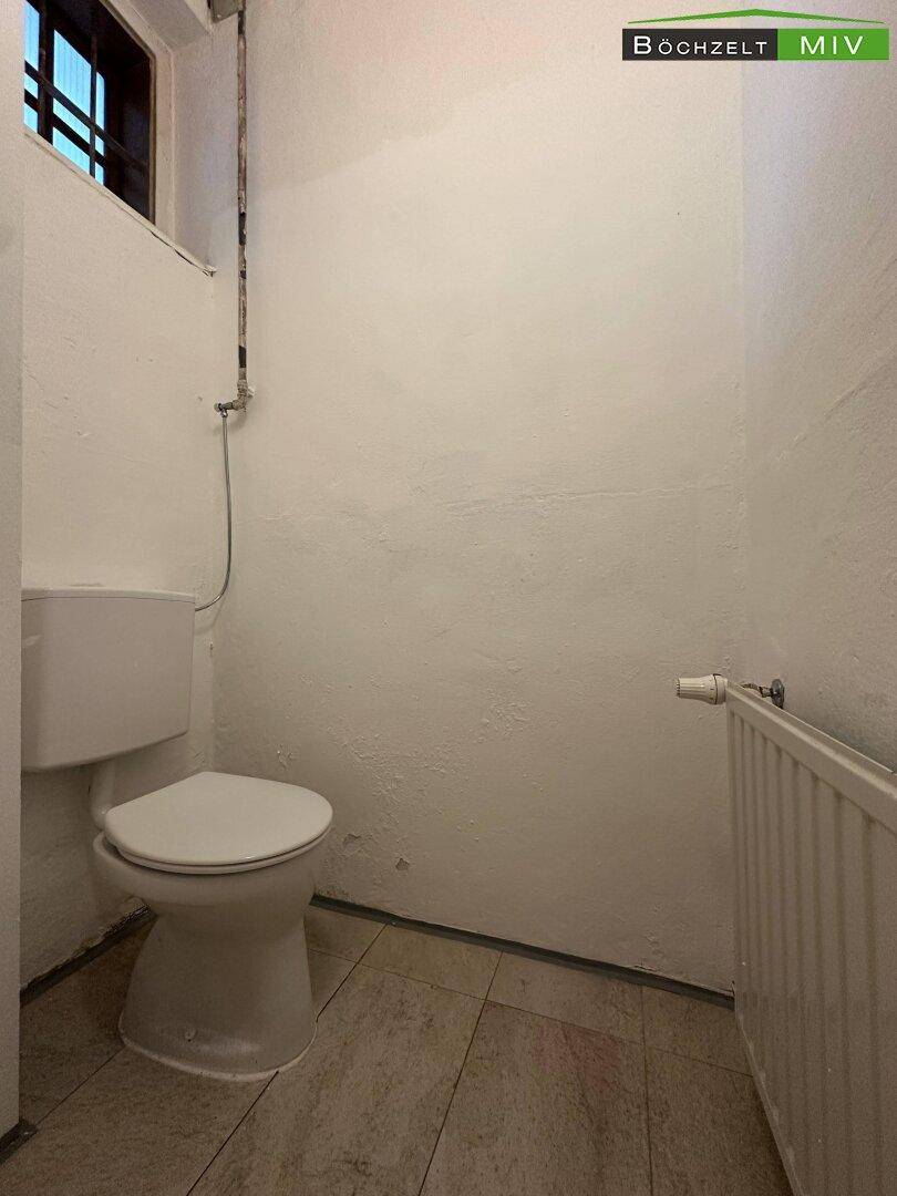 Separate Toilette mit Fenster, Heizkörper und einfachen weißen Fliesen an den Wänden.