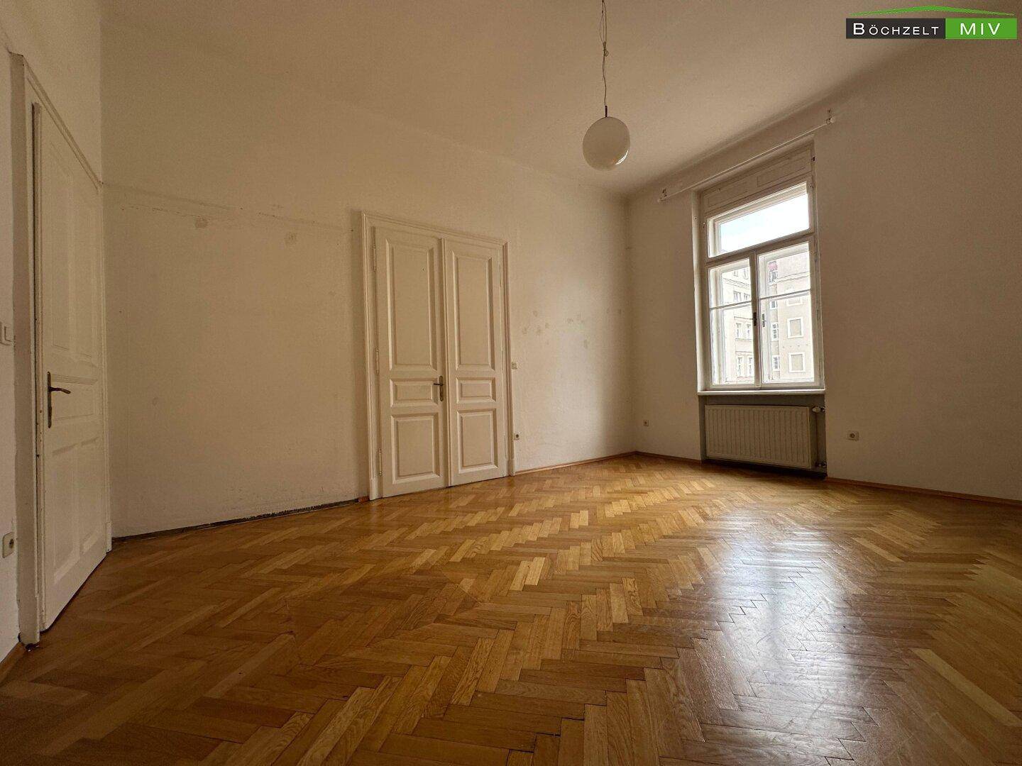 Helles Zimmer mit Fischgrätparkett, hohen Decken und einem großen Fenster, das den Raum erhellt.