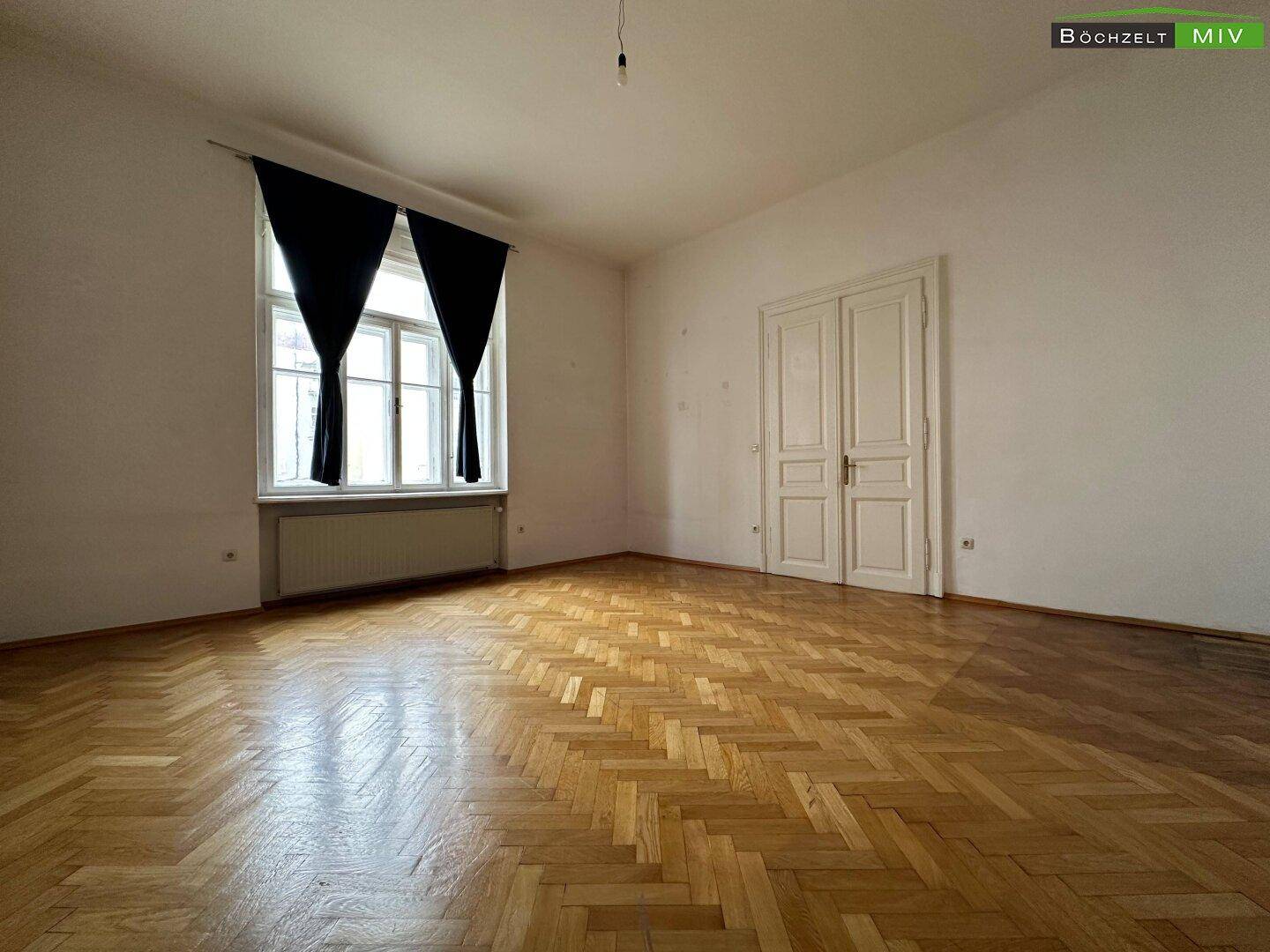 Geräumiges Zimmer mit Fischgrätparkett, hohen Decken und einem großen Fenster mit dunklen Vorhängen.