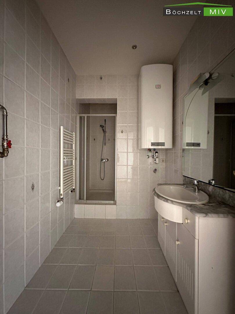 Gefliestes Badezimmer mit Duschkabine, Waschbecken, beheiztem Handtuchhalter und Warmwasserboiler.