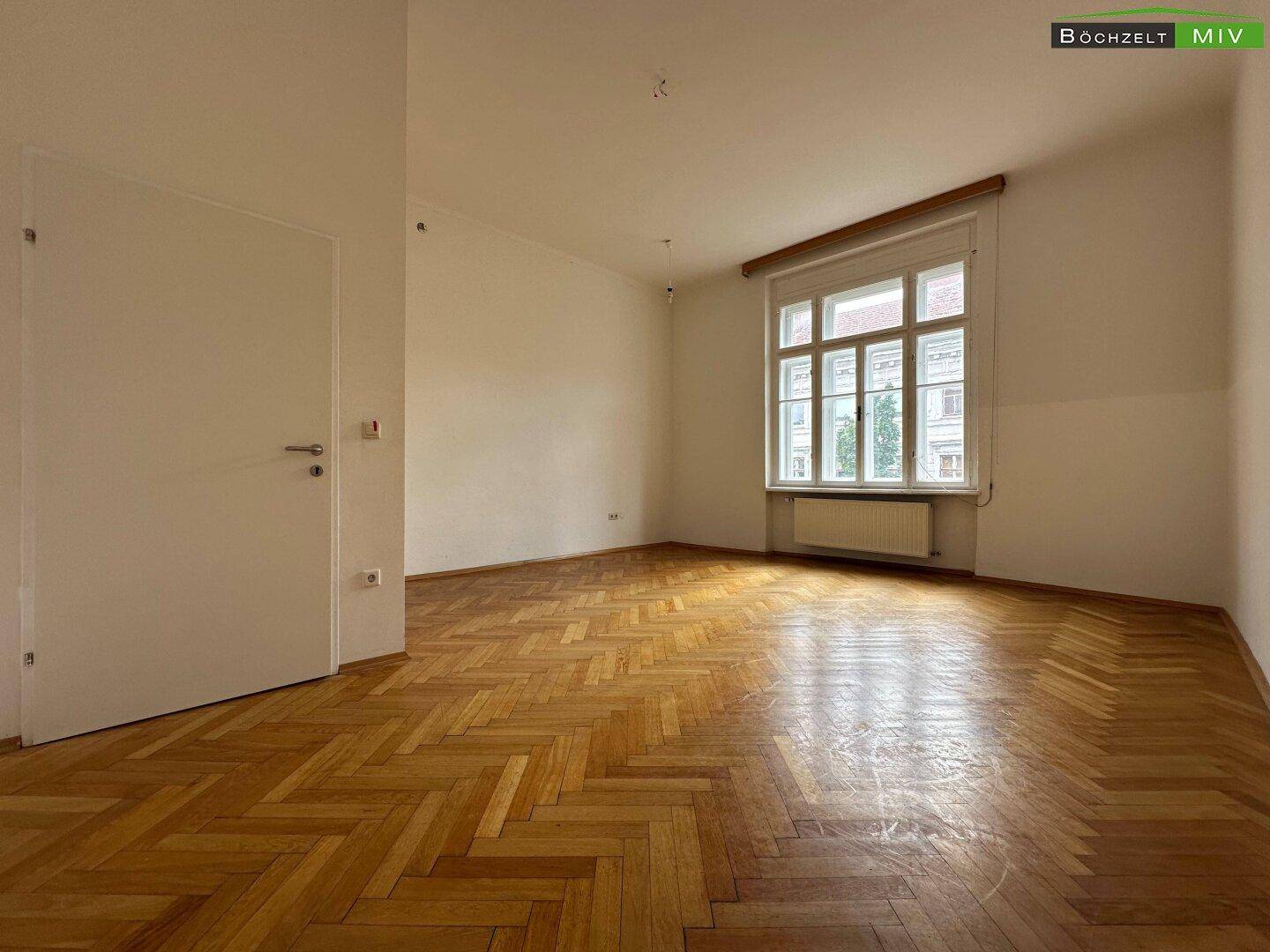 Geräumiges Zimmer mit Fischgrätparkett und einem großen Fenster, das viel Tageslicht hereinlässt.