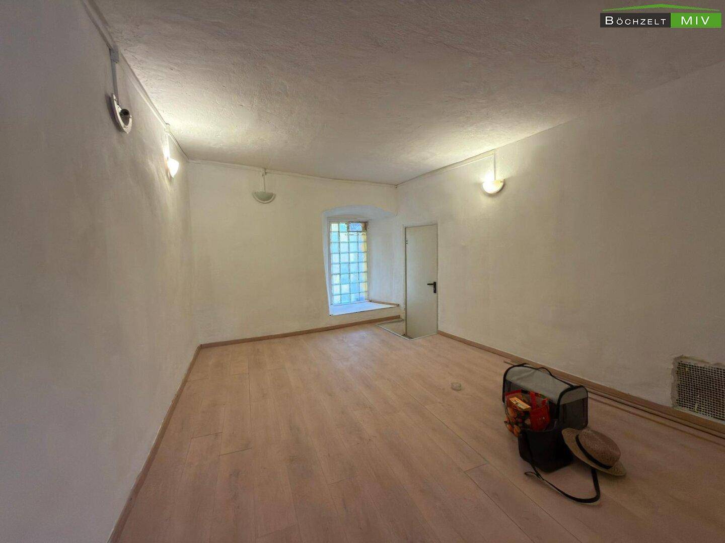 Kleiner Raum mit Holzboden, einer Tür und einem Fenster aus Glasbausteinen, beleuchtet von Wandlampen.