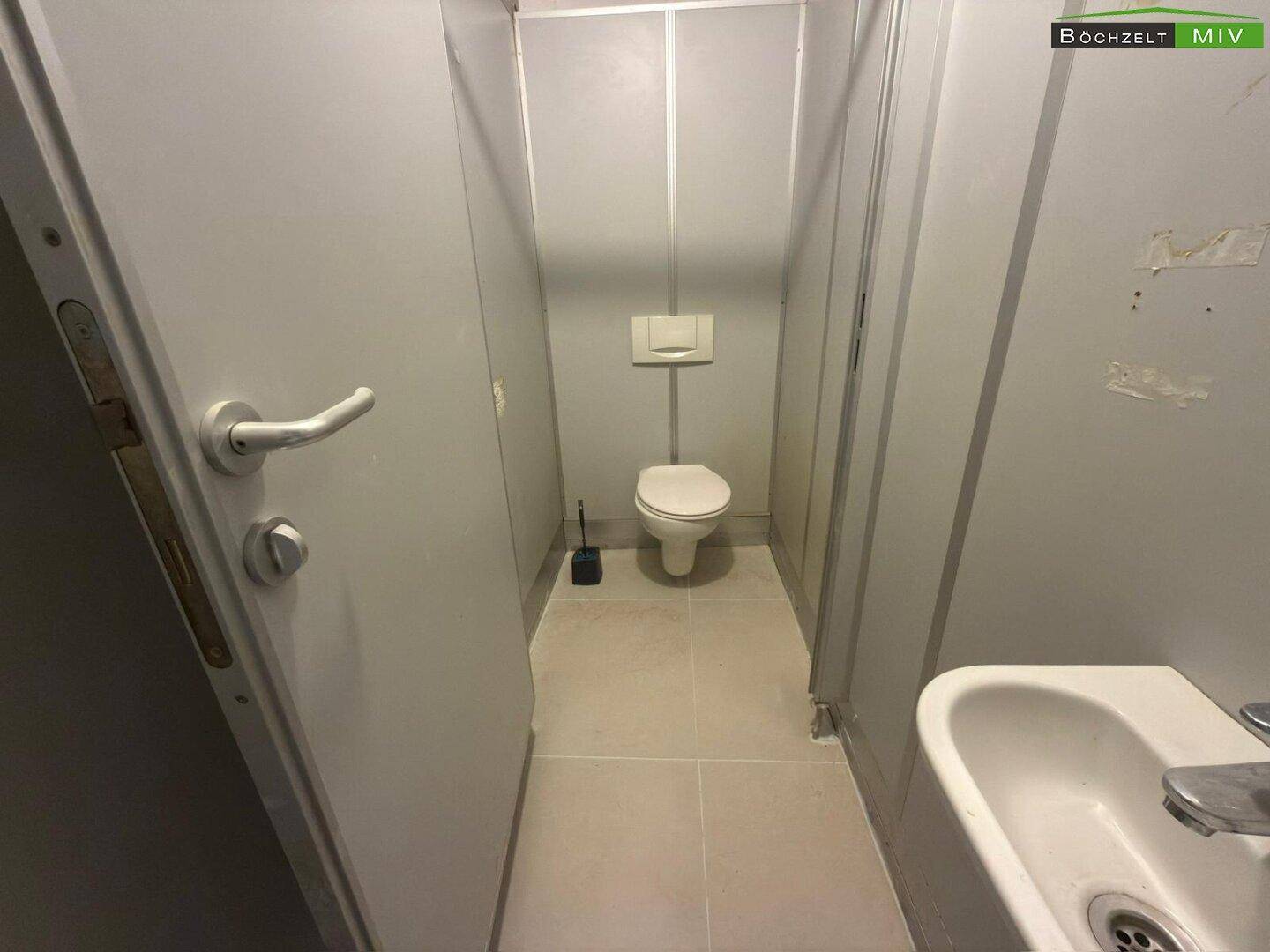 Schmale Toilettenkabine mit Wand-WC und kleinem Waschbecken im Vordergrund.