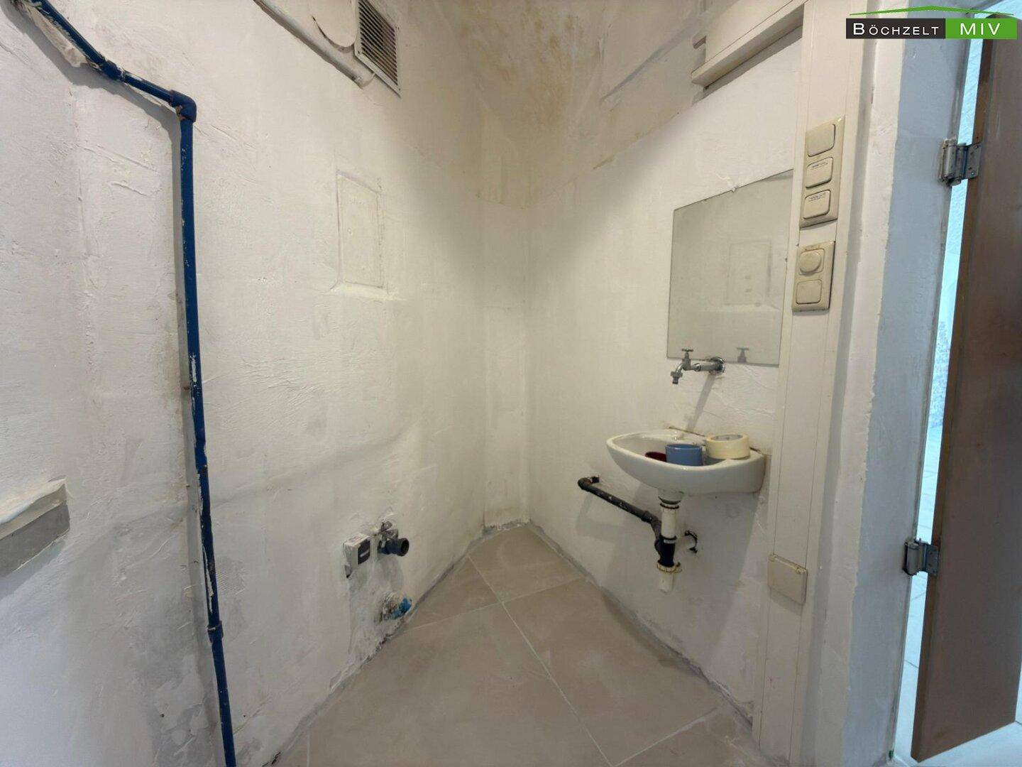 Einfacher Waschraum mit Waschbecken, Spiegel und sichtbaren Wasserleitungen an der Wand.