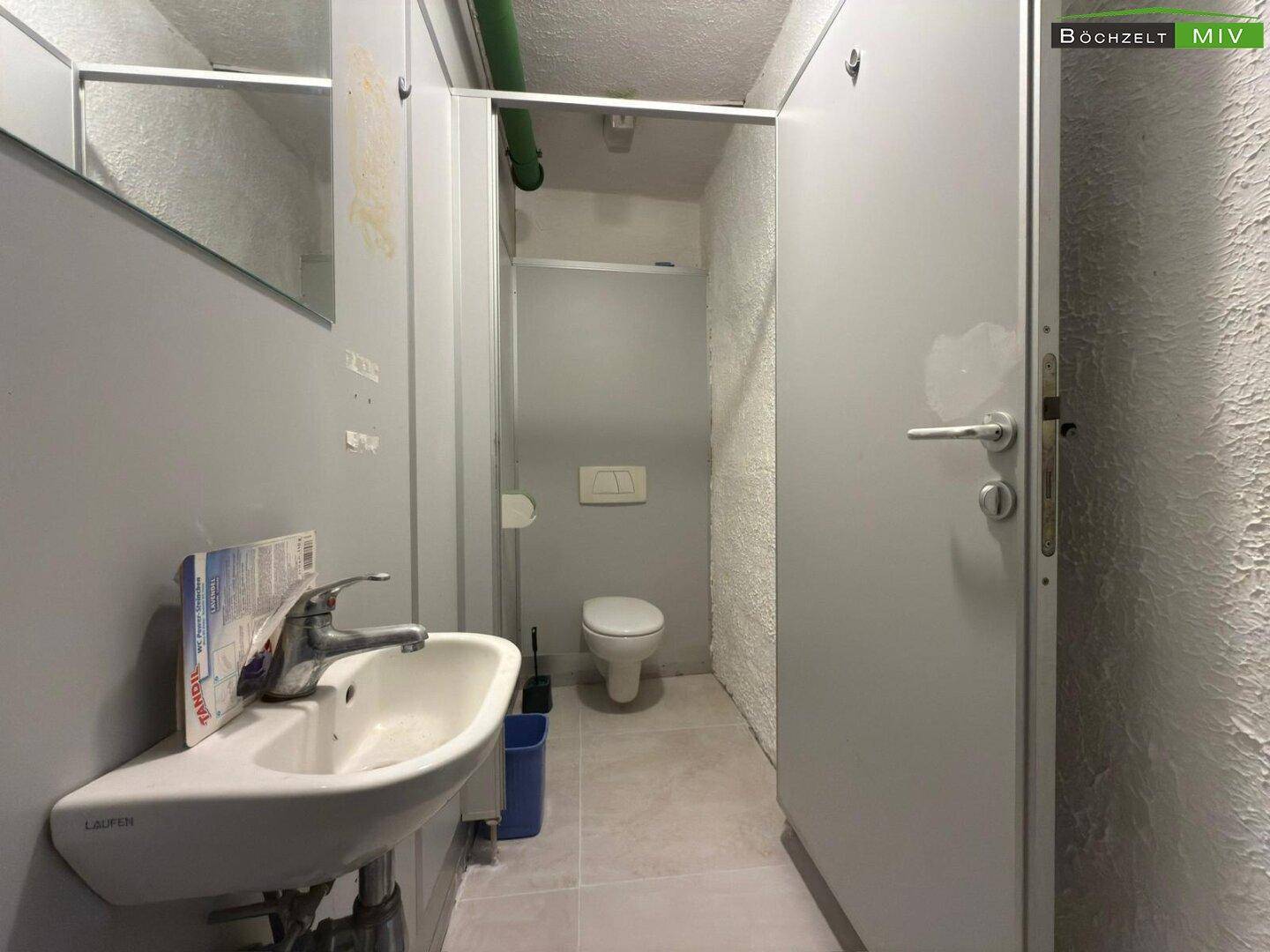 Toilettenbereich mit Waschbecken, Spiegel und einer weiteren Kabine mit Wand-WC.