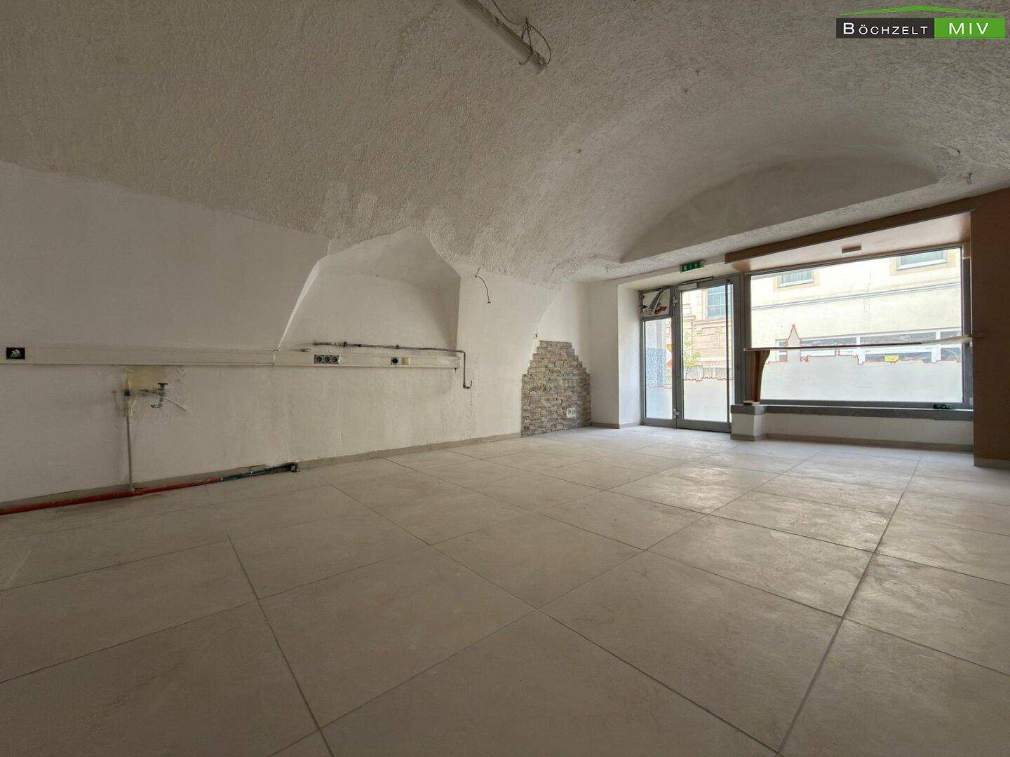 Großer, leerer Raum mit gewölbter Decke, Fliesenboden und großen Fenstern zur Straße hin.