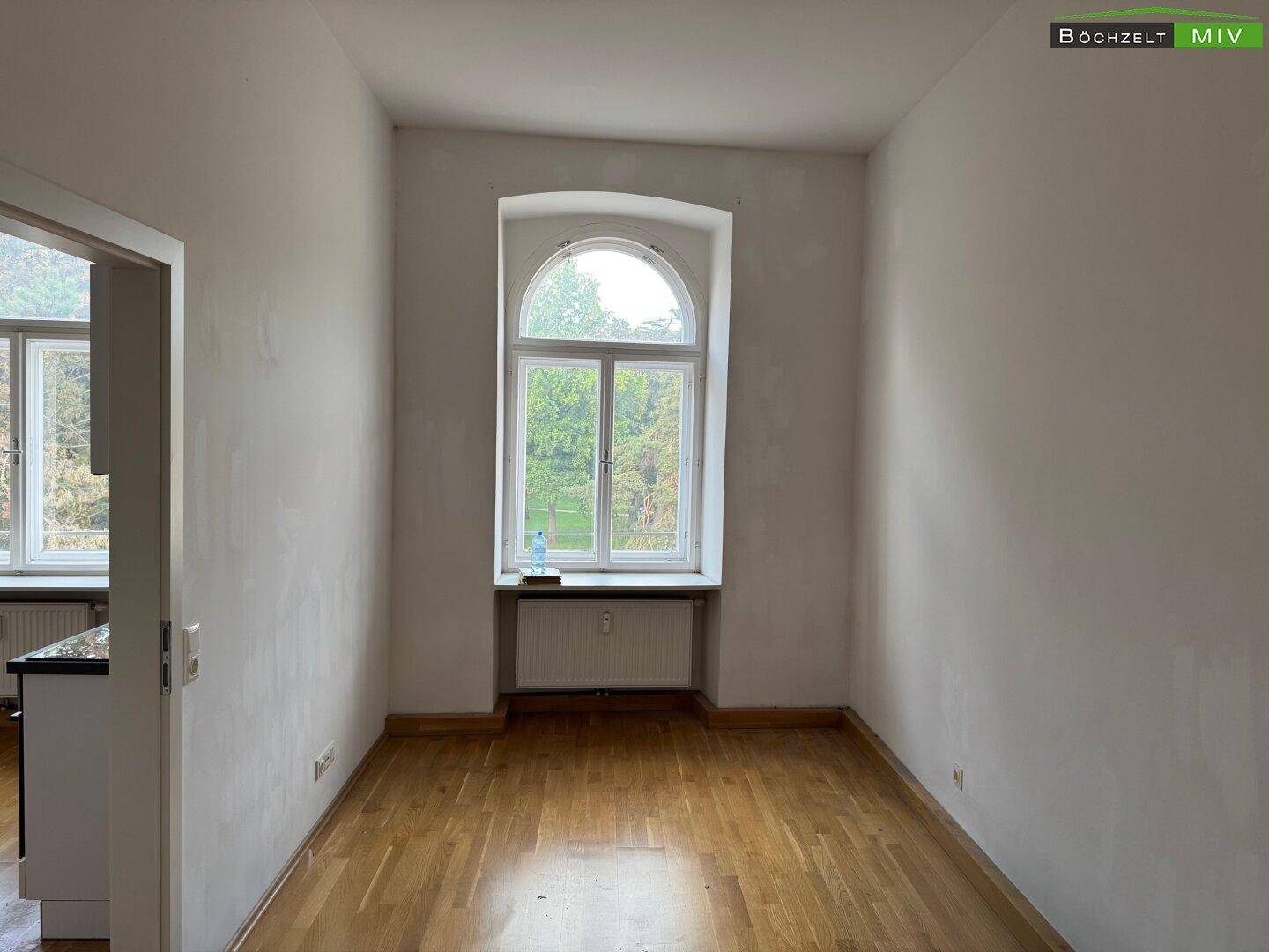Leeres Zimmer mit elegantem Rundbogenfenster, Holzboden und Blick ins Grüne.