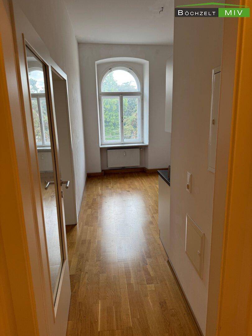 Flur mit Holzboden, der in ein helles Zimmer mit Rundbogenfenster und angrenzender Küchenzeile führt.