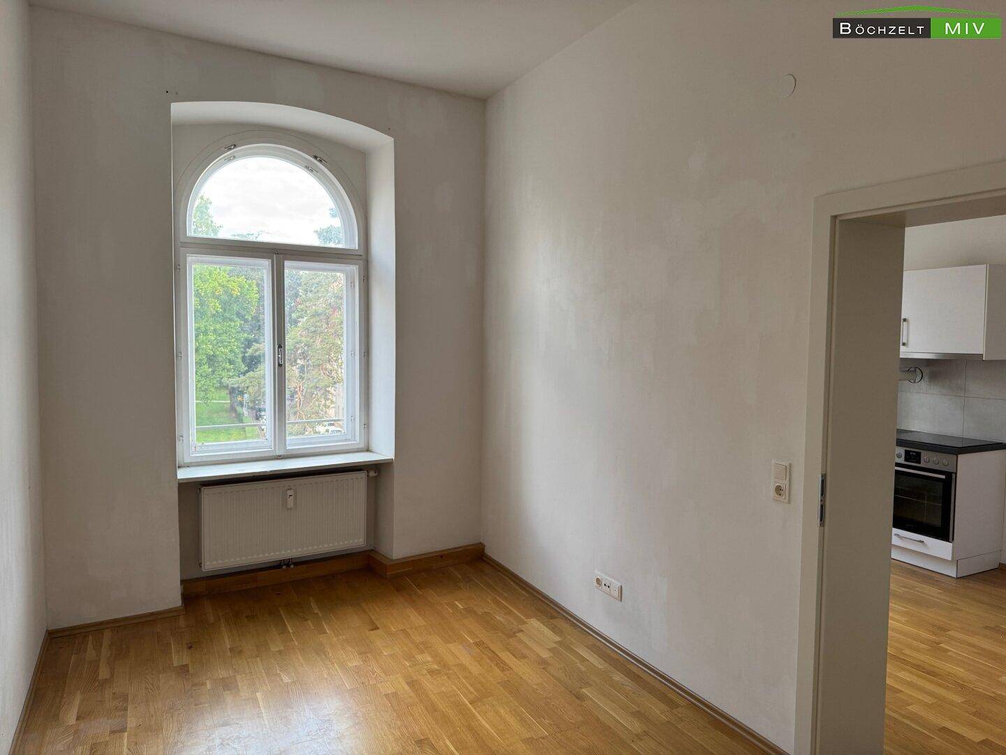 Helles Zimmer mit Holzboden und großem Rundbogenfenster, angrenzend an die Küchenzeile.
