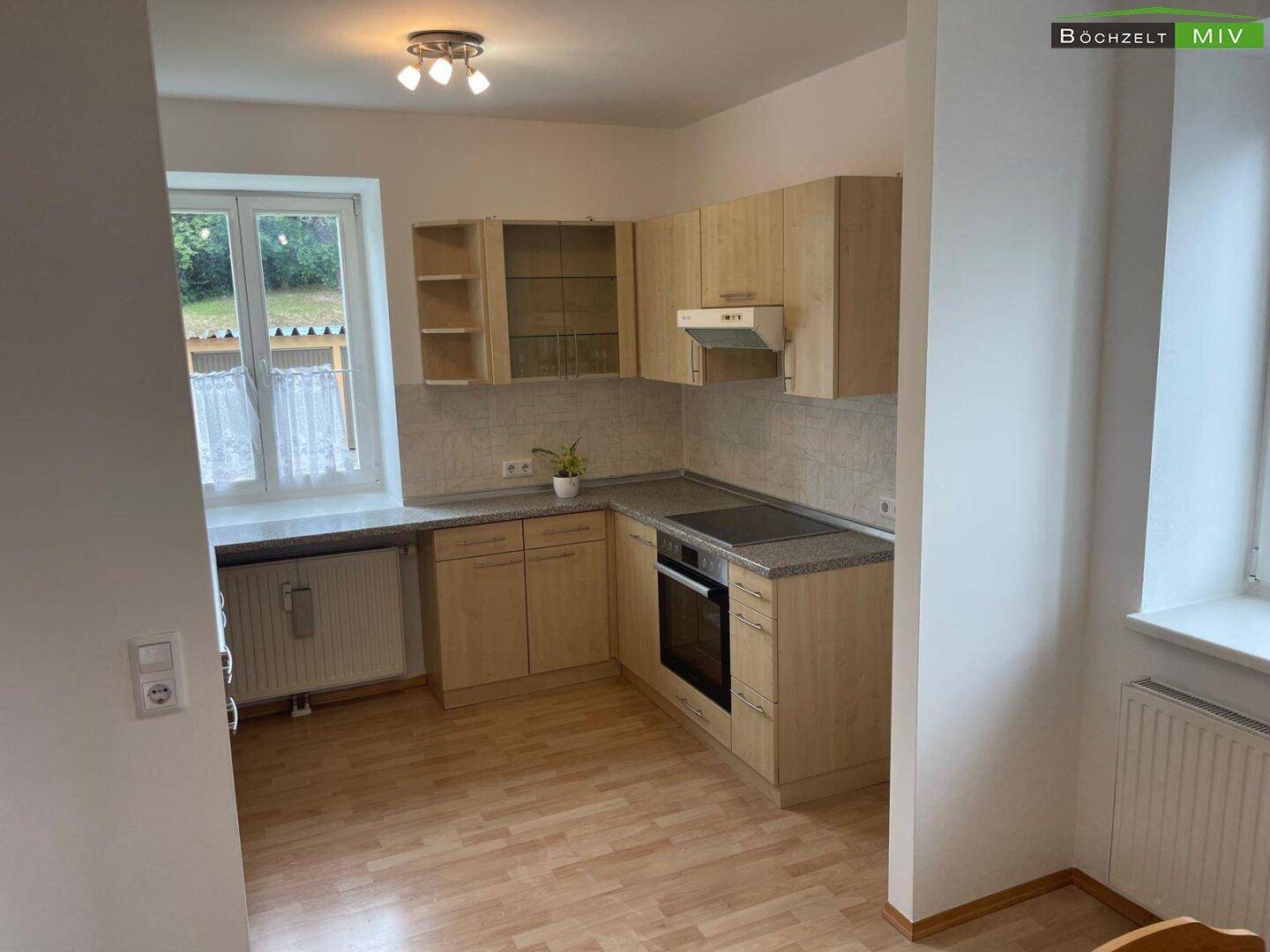 Funktionale Küche mit hellen Holzschränken, grauer Arbeitsplatte und Fenster mit Blick ins Grüne.
