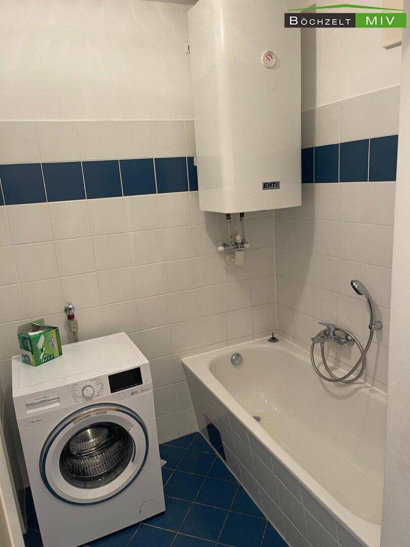 Badezimmer mit Badewanne, Waschmaschine und Warmwasserboiler, gefliest in Weiß und Blau.