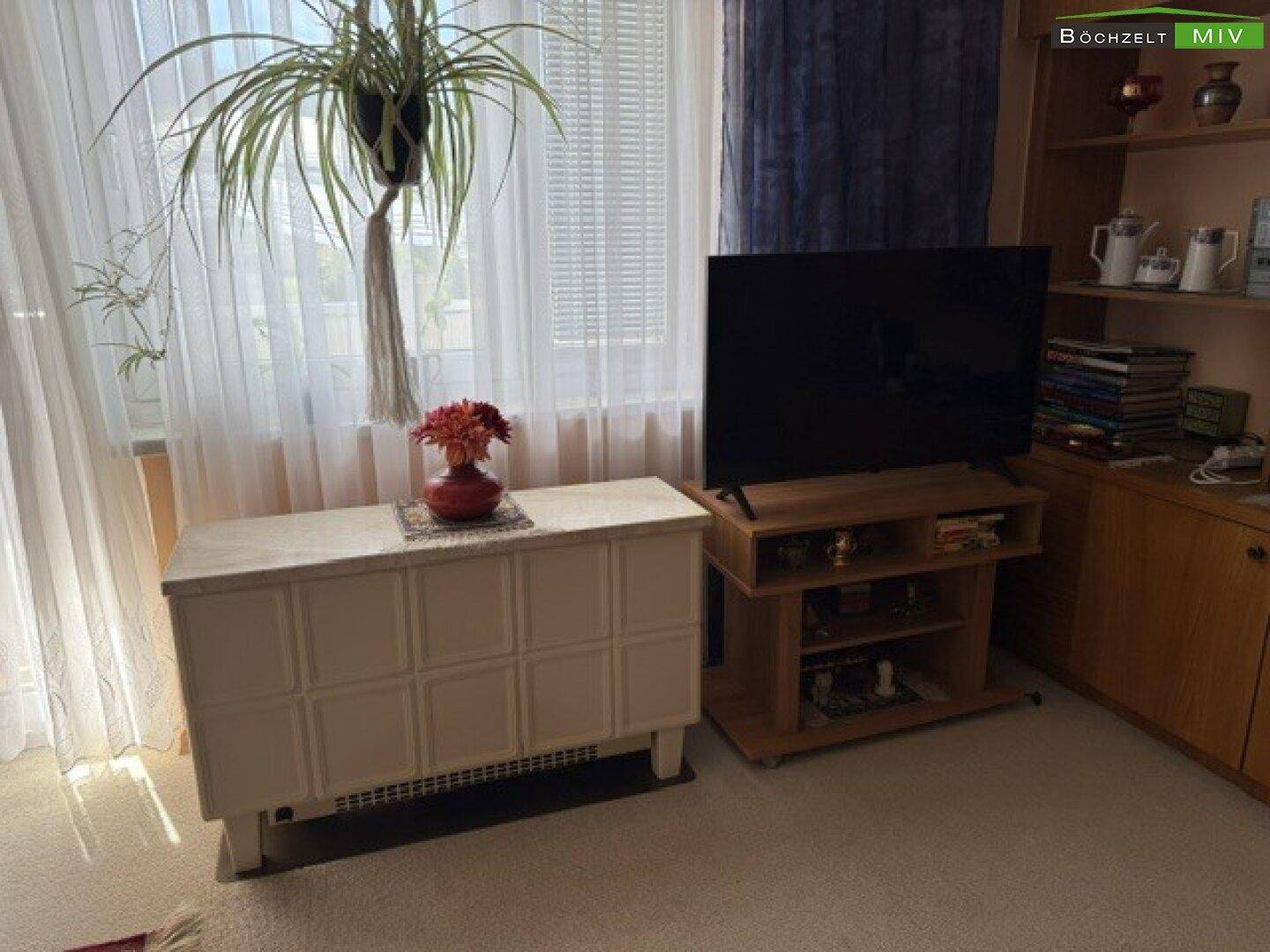 Helles Wohnzimmer mit Teppichboden, großem Fenster und einem Kachelofen.