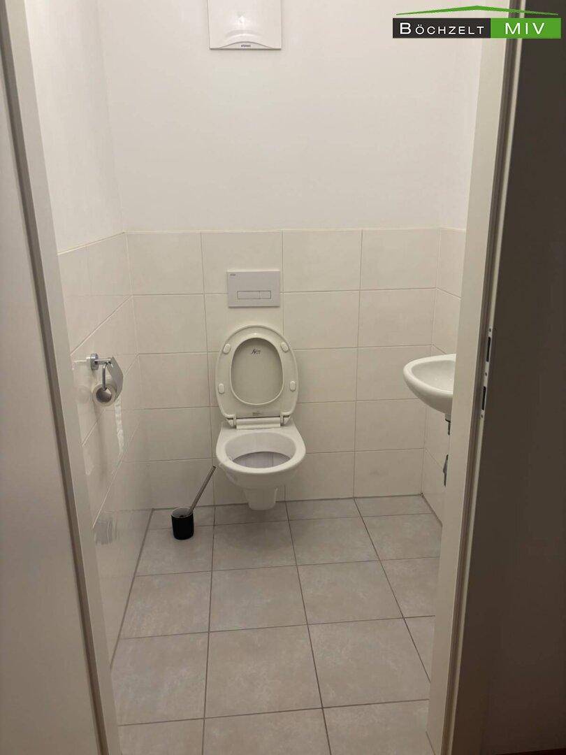 Separates WC mit Toilette, kleinem Waschbecken und hellen Fliesen, sauber und funktional.