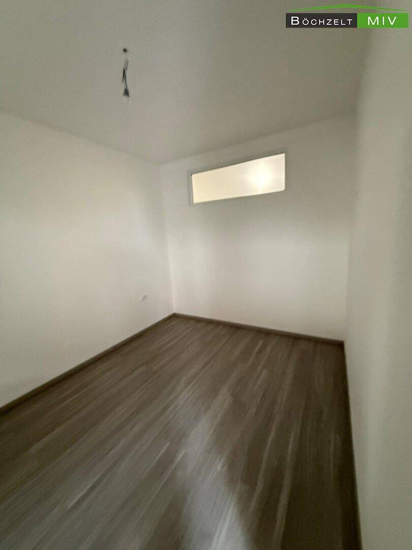 Helles, leeres Zimmer mit Laminatboden und einem hohen Fenster für Tageslicht.