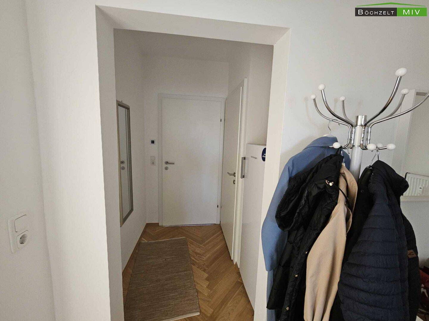Schmaler Flur mit Fischgrätparkett, Garderobe und weißer Eingangstür.