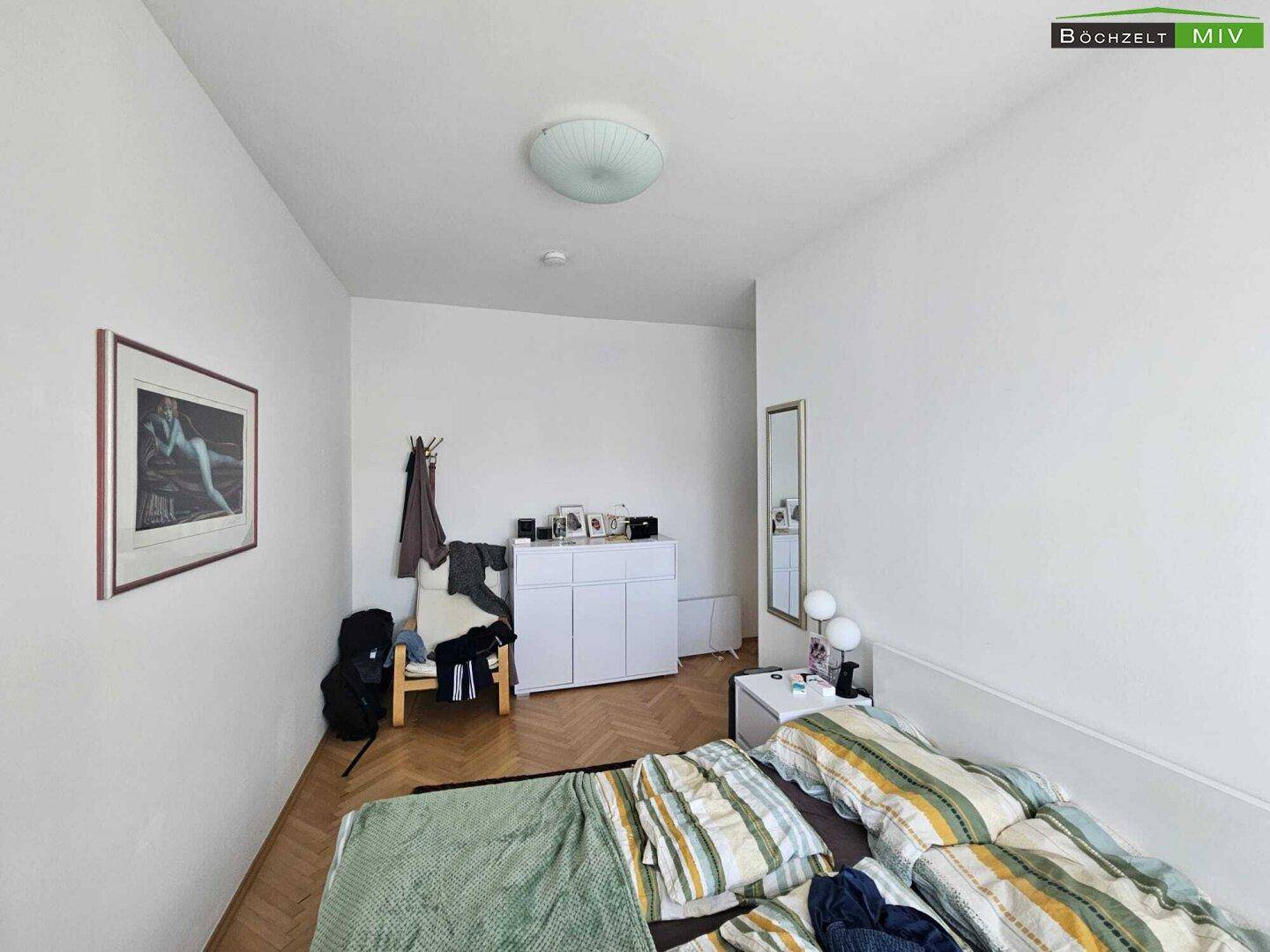 Schlafzimmer mit Bett, Kommode, Spiegel und Fischgrätparkettboden.